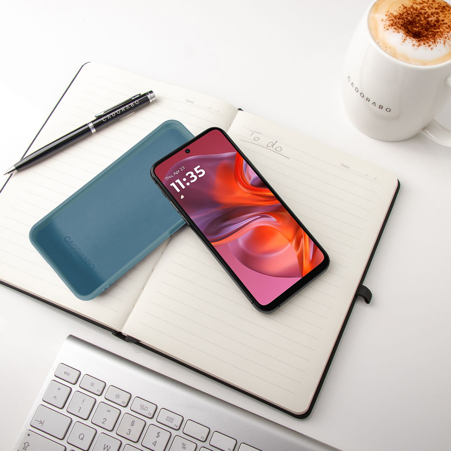 Ein Smartphone, Notizbuch, Stift, Tastatur und Kaffee liegen auf einem weißen Tisch.