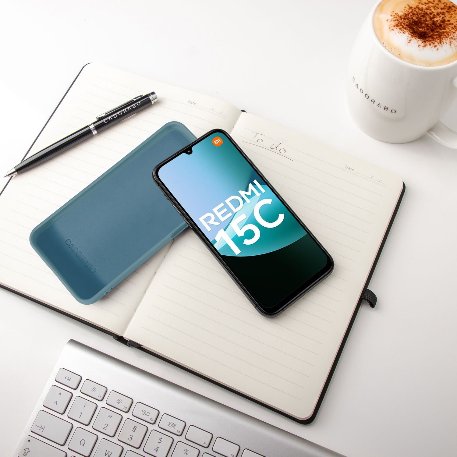 Ein Redmi 15C Handy, blaue Hülle, Stift, Notizbuch, Kaffee und Tastatur auf einem weißen Schreibtisch.