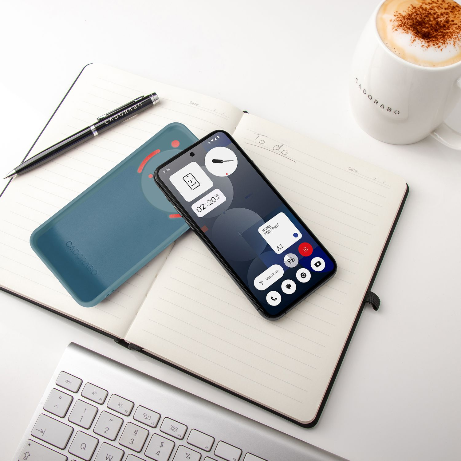 Ein Telefon, blaue Hülle, Stift, Tastatur und Kaffee liegen auf einem Notizbuch.