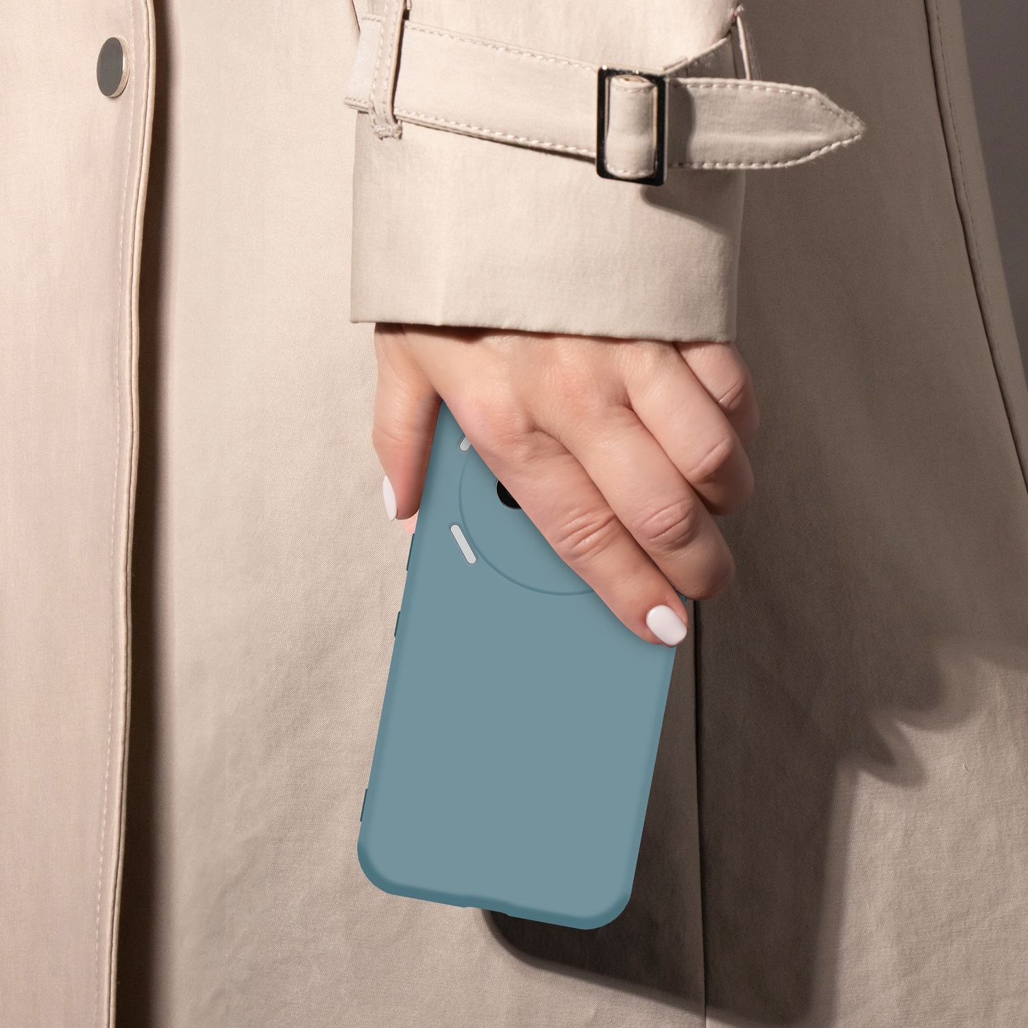 Eine Hand mit weißen Nägeln hält eine teal Handyhülle vor einem hellbraunen Trenchcoat.