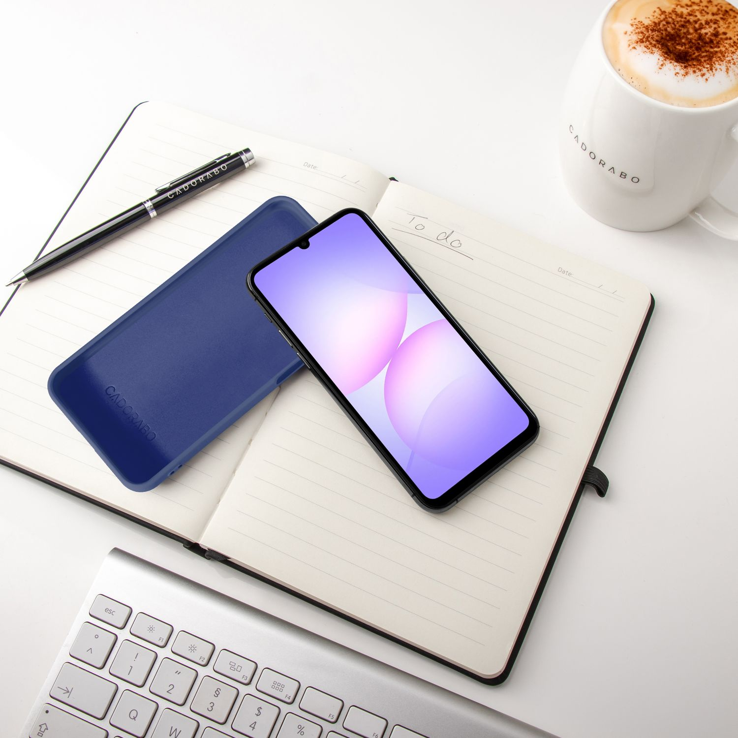 Ein Notizbuch mit Handy, Stift, Tastatur und Kaffeetasse auf weißem Schreibtisch.