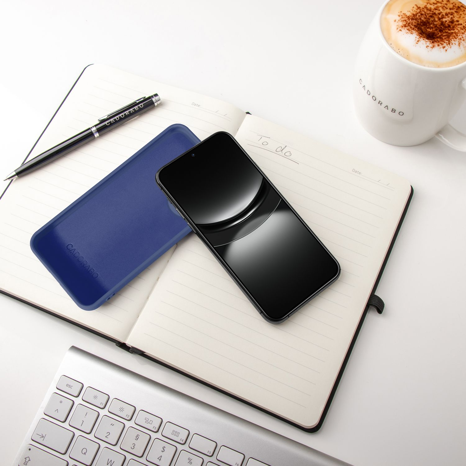 Ein aufgeschlagenes Notizbuch mit Stift, Handy, blauem Etui, Tastatur und Kaffee.