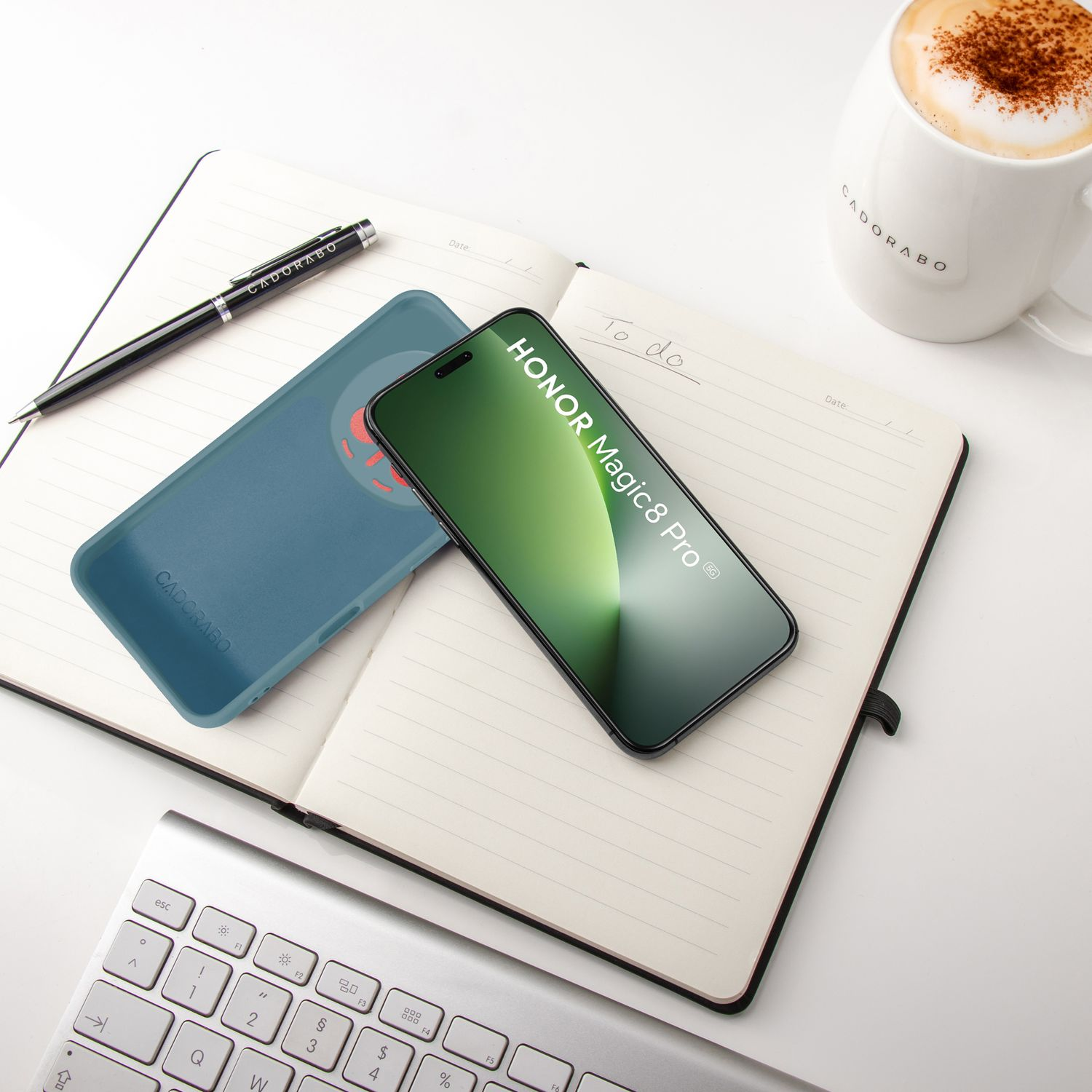 Ein Handy, Notizbuch, Stift, Tastatur und eine Kaffeetasse auf einem weißen Tisch.