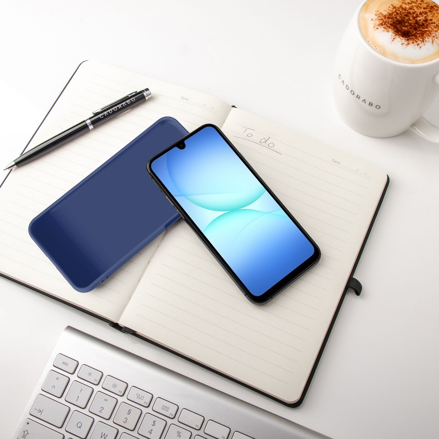 Ein Notizbuch mit Handy, Stift, Tastatur und Kaffeetasse auf weißer Fläche.