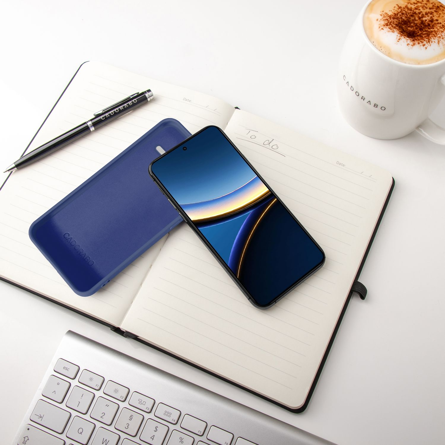 Ein Smartphone liegt auf einem Notizbuch neben einer blauen Hülle, einer Tasse Kaffee und einer Tastatur.