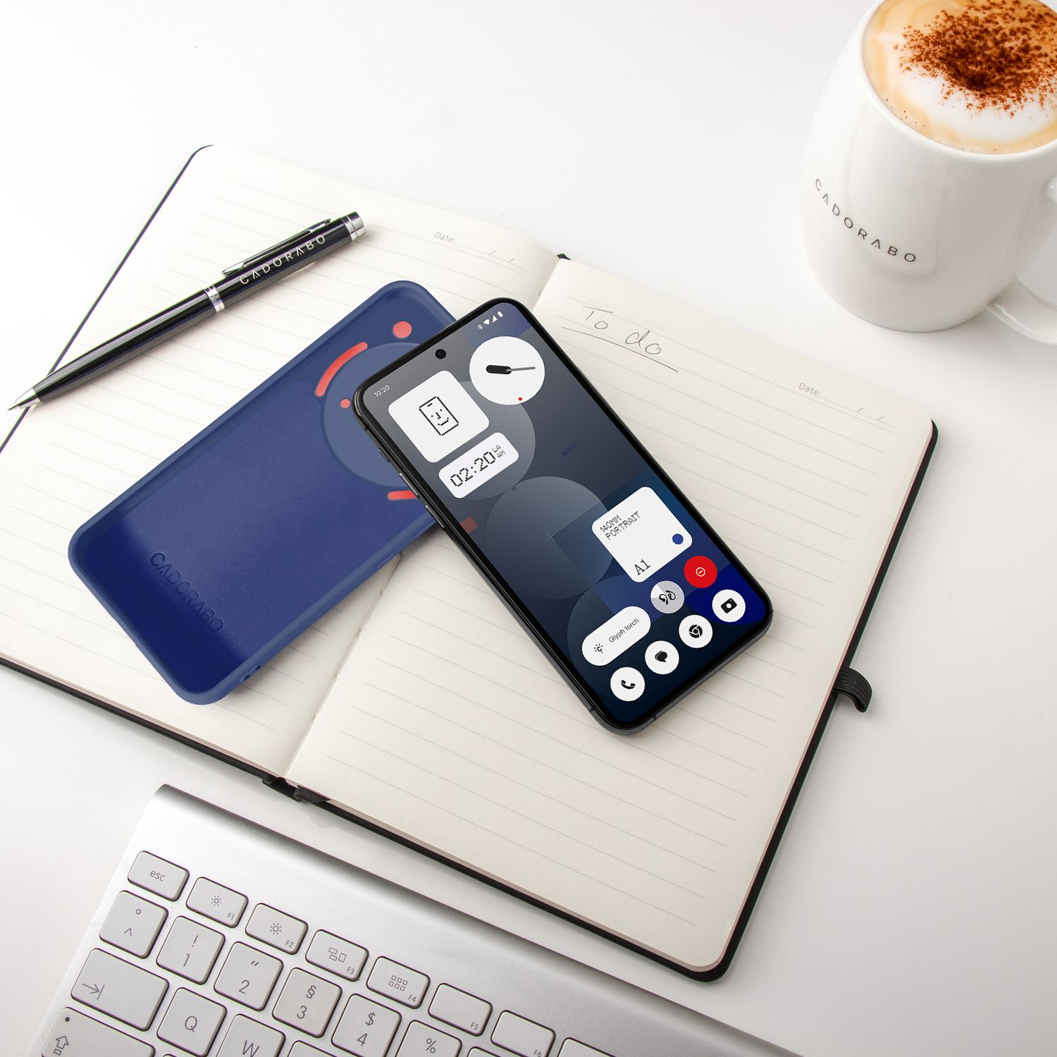 Smartphone, blaue Hülle, Stift, Notizbuch, Tastatur und Kaffeetasse auf weißer Fläche.