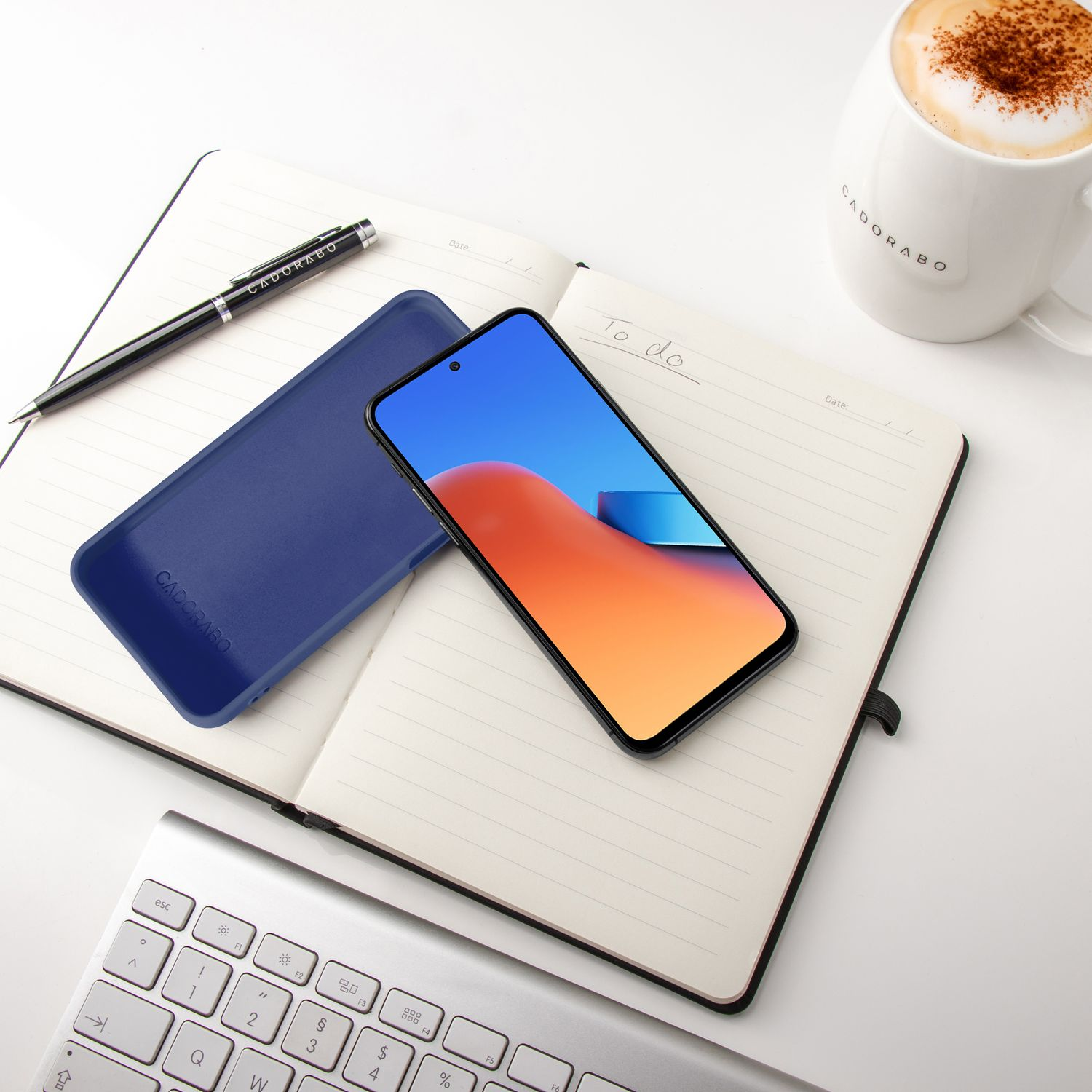 Ein Smartphone liegt auf einem Notizbuch neben einer blauen Hülle, Tastatur und einer Tasse Kaffee.