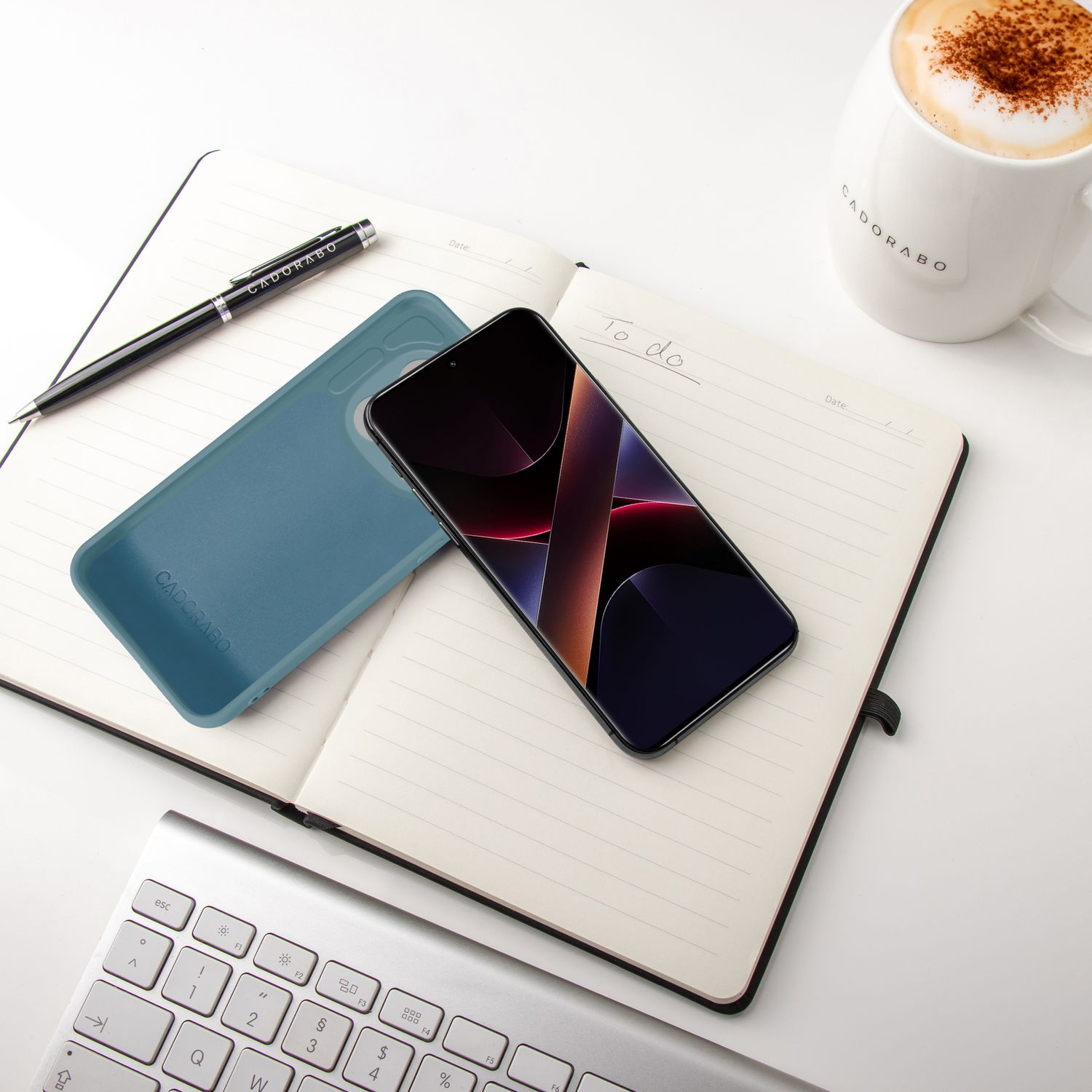 Ein Smartphone, Notizbuch, Stift, Tastatur und eine Tasse Kaffee liegen auf einem weißen Tisch.