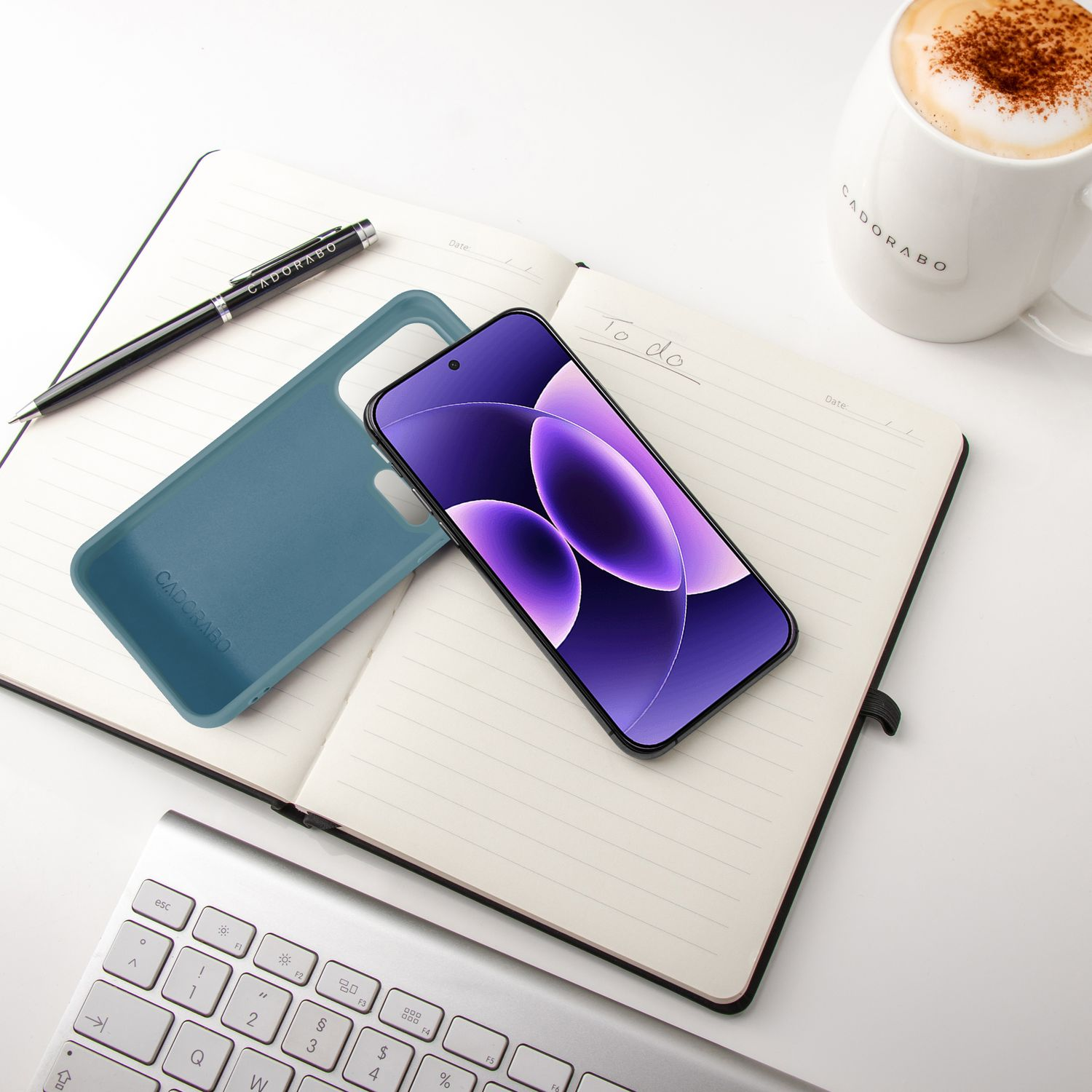 Ein Smartphone, Hülle, Notizbuch, Stift, Tasse Kaffee und Tastatur auf weißem Tisch.