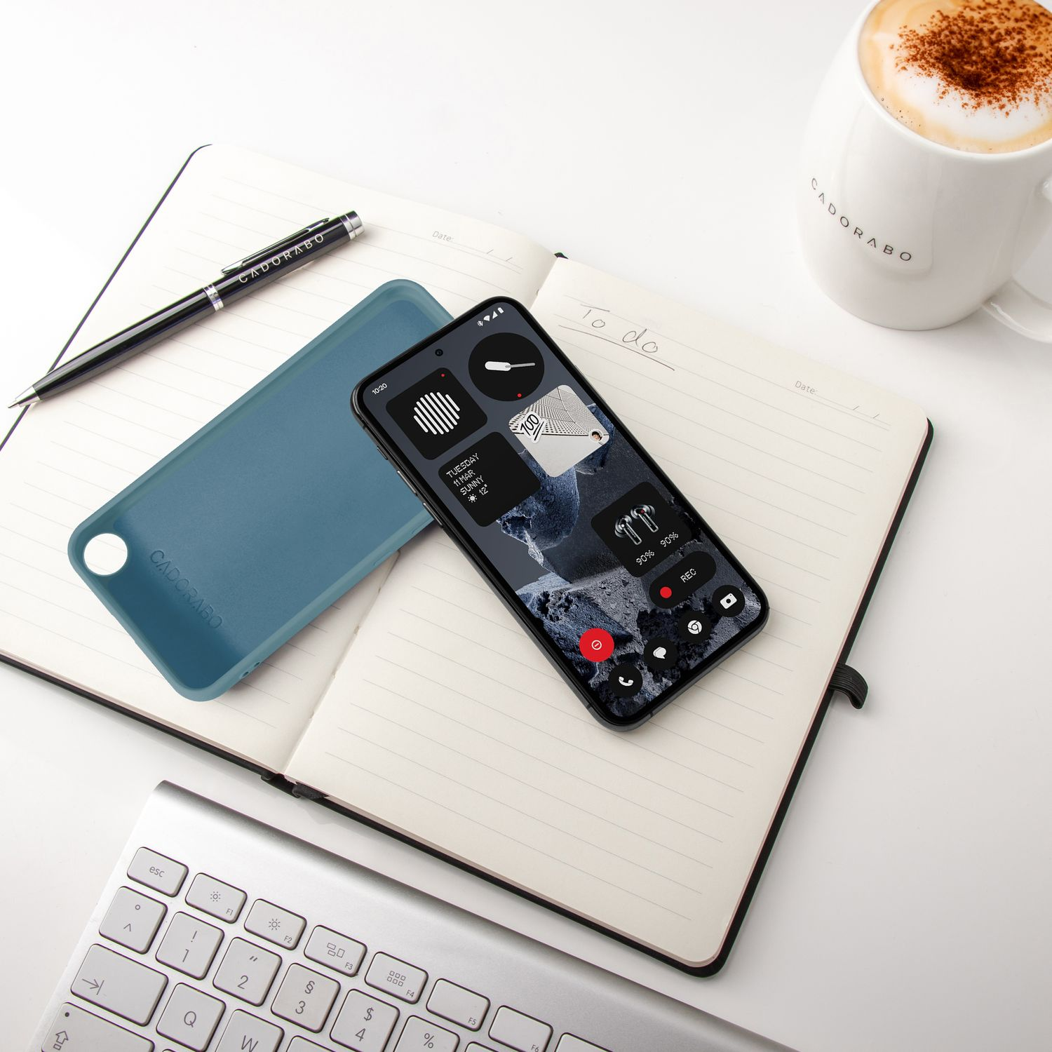 Ein Smartphone auf einem Notizbuch, daneben eine blaue Handyhülle, Stift, Tasse Kaffee und Tastatur.