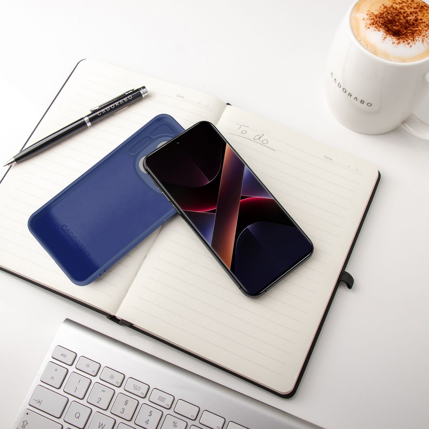 Ein Smartphone auf einem Notizbuch, daneben eine blaue Hülle, Stift, Kaffee und Tastatur.