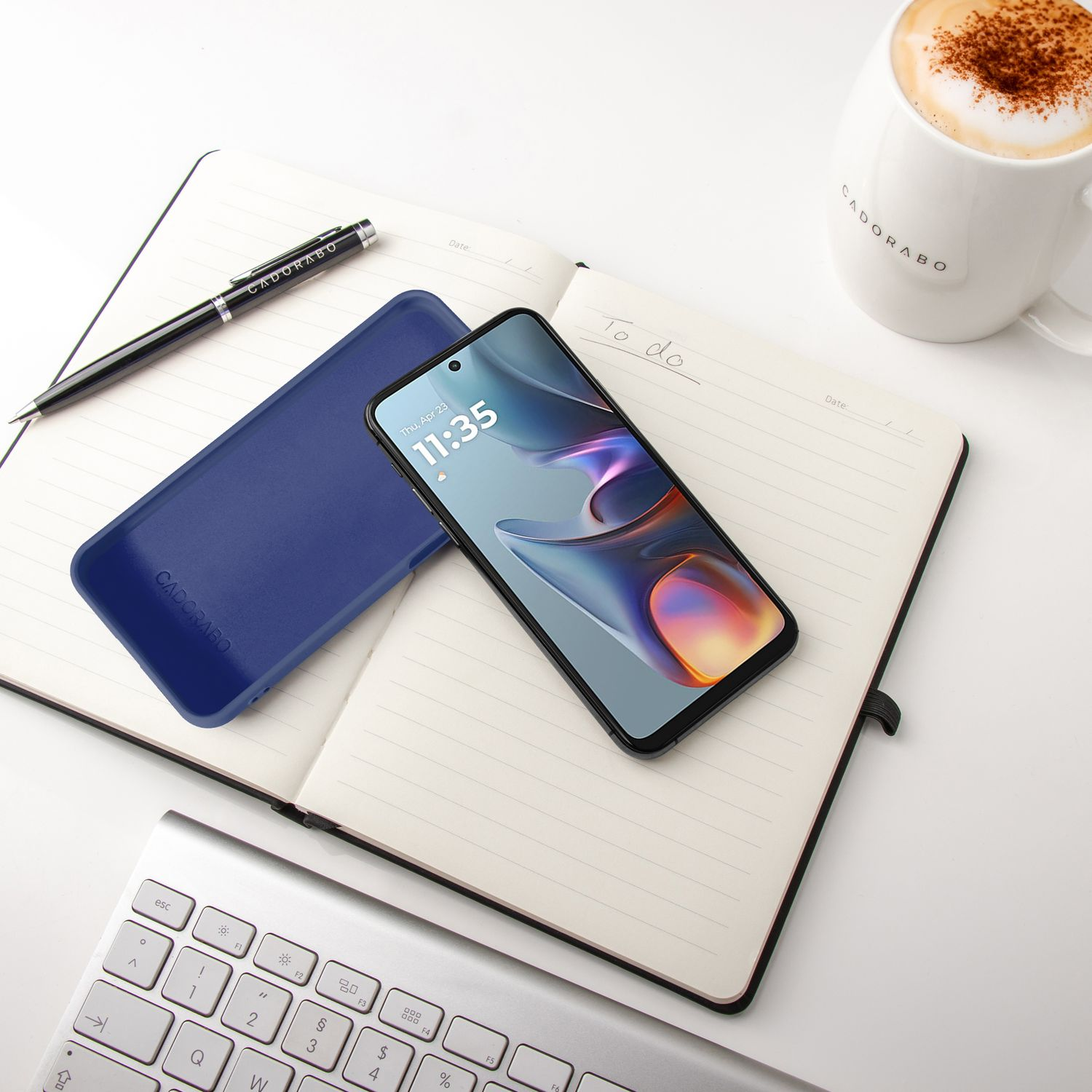 Ein Handy, Notizbuch, Stift, Tastatur und eine Kaffeetasse liegen auf einem weißen Tisch.