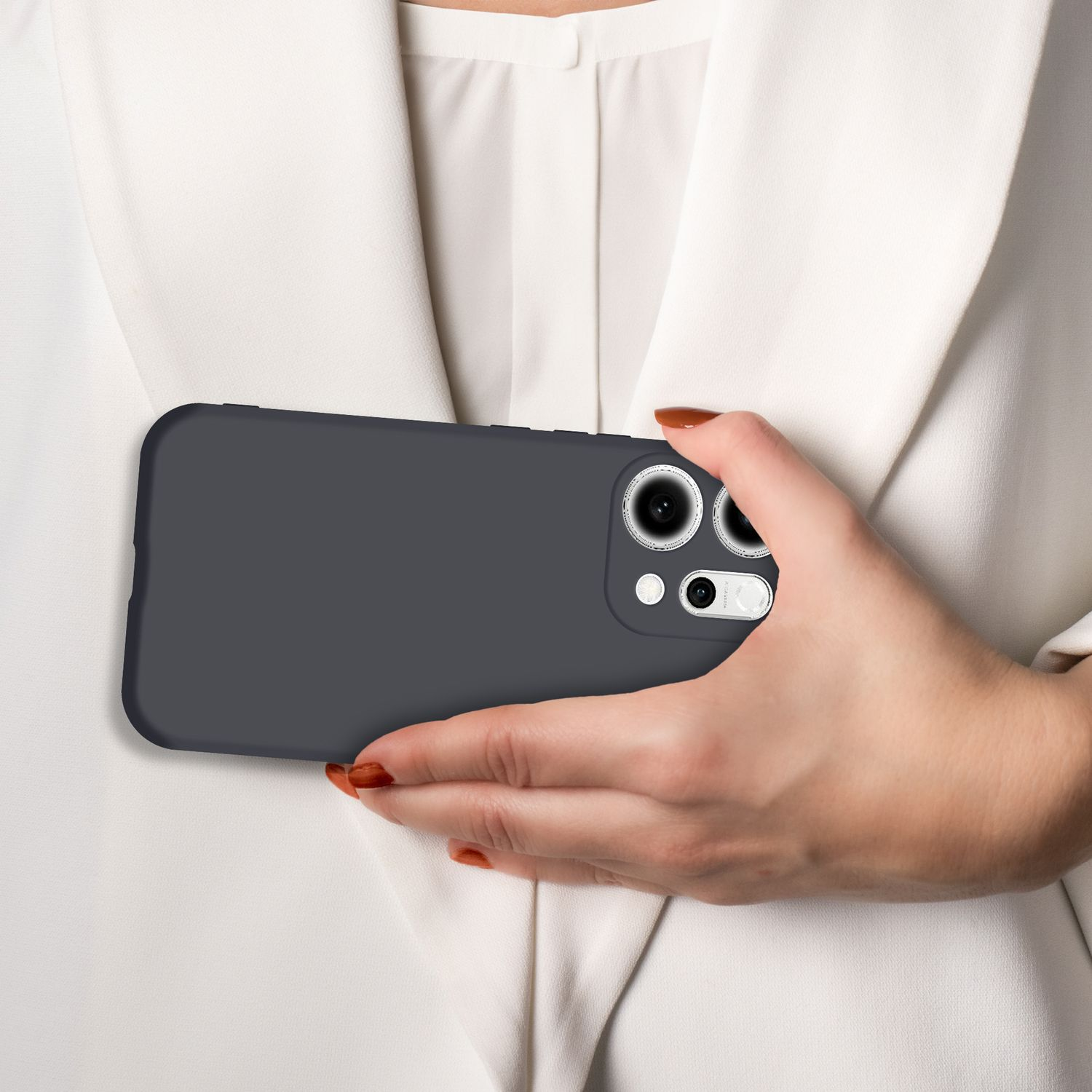 Eine Hand mit braun lackierten Nägeln hält ein dunkelgraues Handy mit Kamera vor einem weißen Blazer.