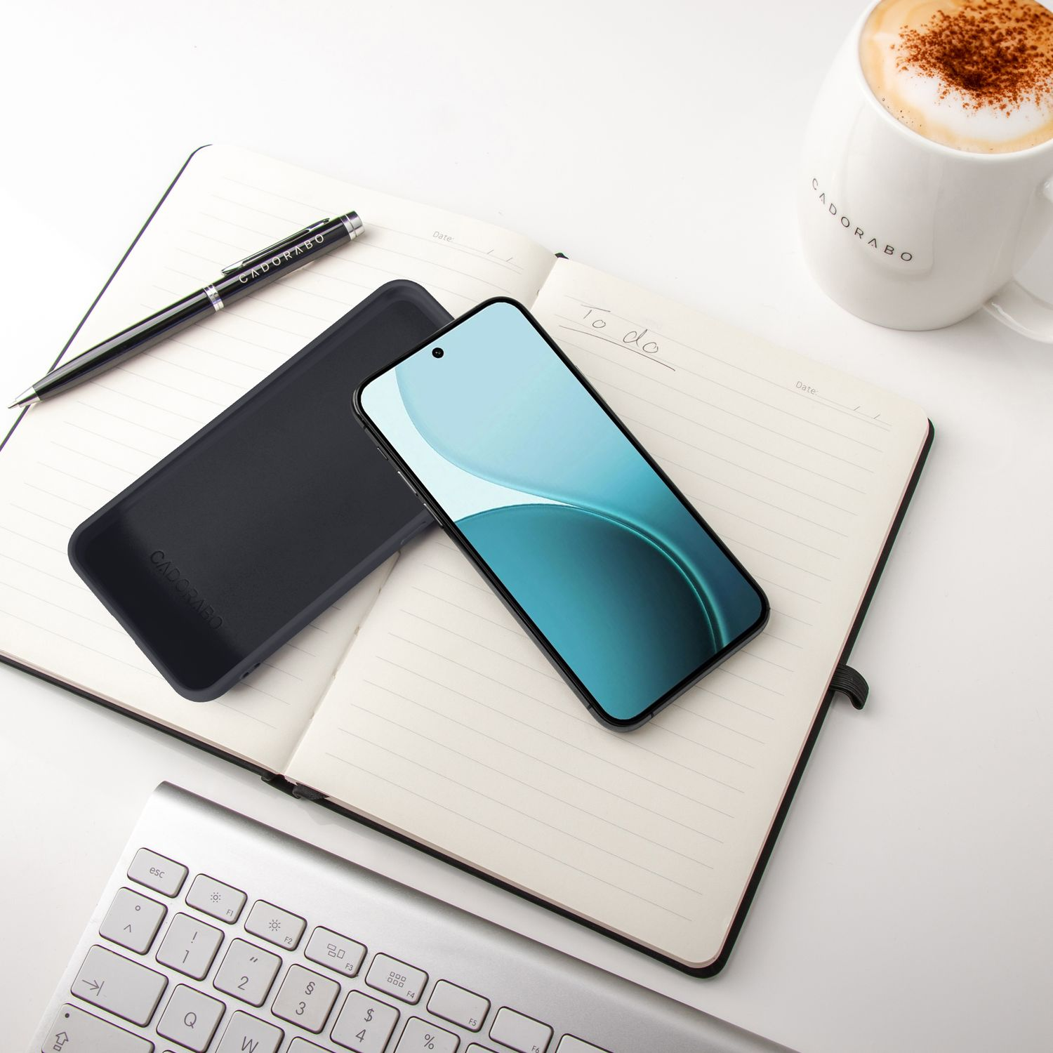 Ein Handy, Notizbuch, Stift, Tasse Kaffee und Tastatur auf einem weißen Schreibtisch.