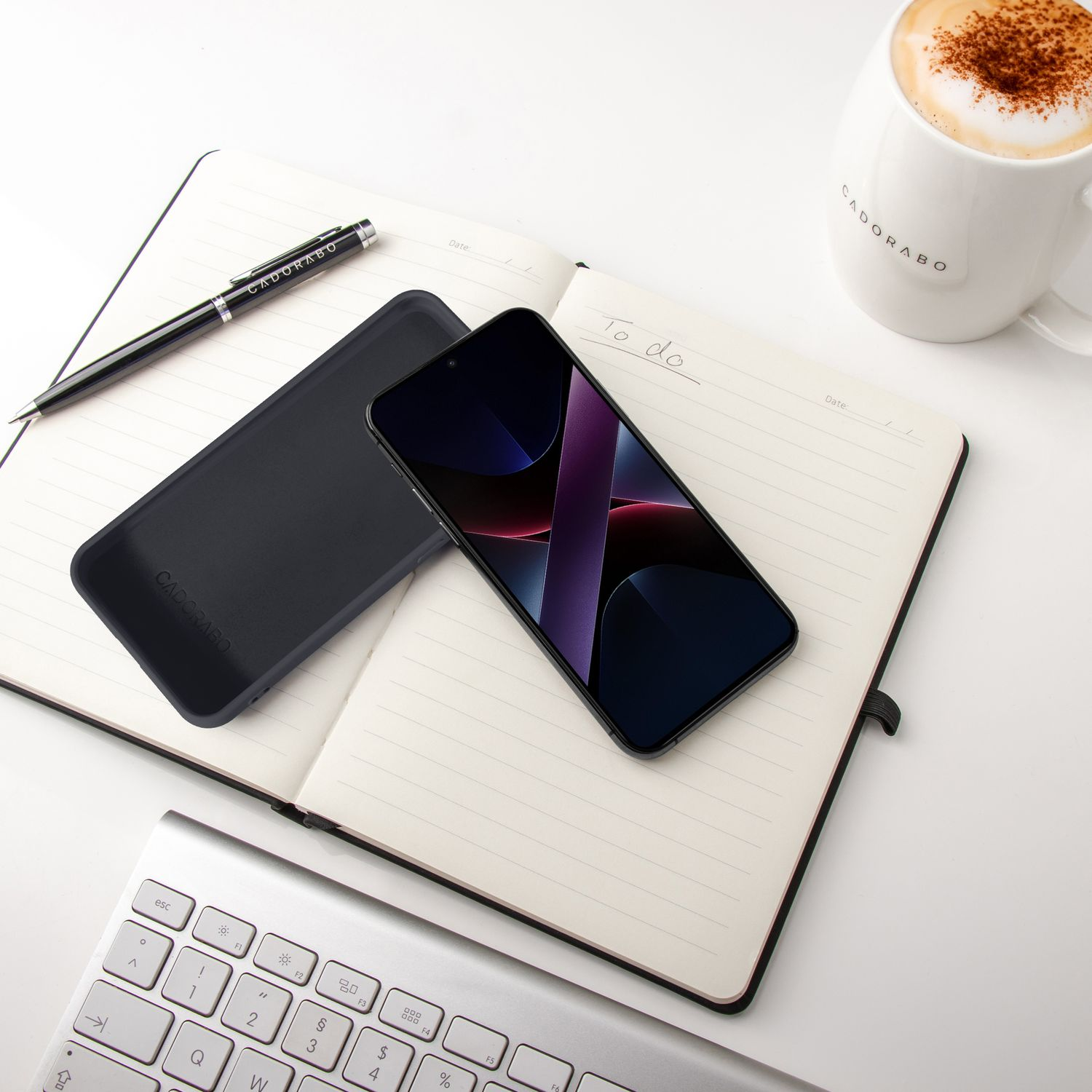 Schwarzes Smartphone auf Notizbuch neben Tastatur und Kaffeebecher.