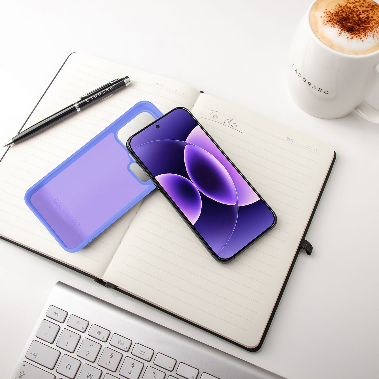 Ein Smartphone, eine lilafarbene Hülle, ein Notizbuch, ein Stift, eine Tastatur und eine Kaffeetasse.