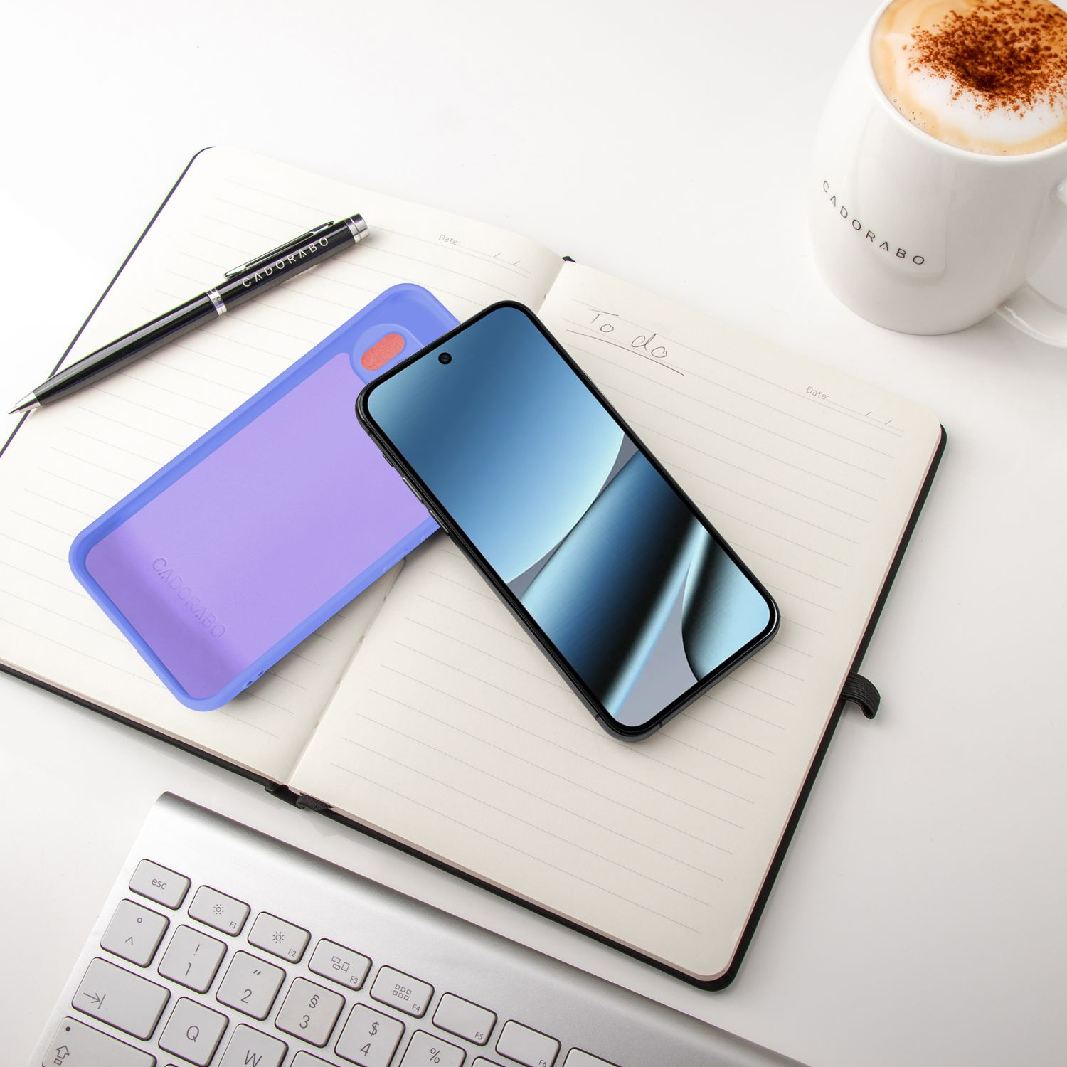 Ein Notizbuch, Handy, lila Hülle, Stift, Tastatur und Kaffeetasse auf weißem Untergrund.