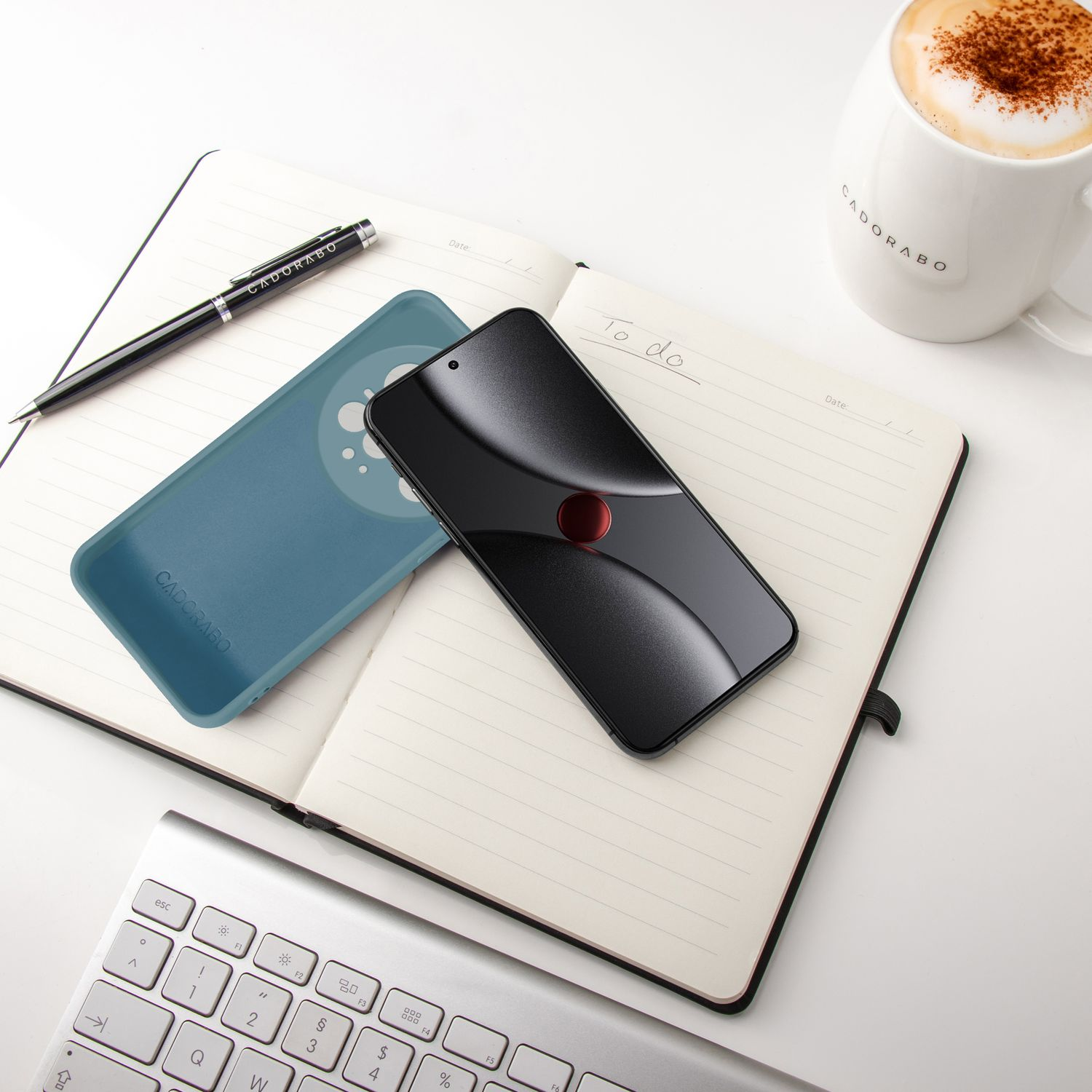 Ein schwarzes Smartphone, eine blaue Hülle, Notizbuch, Stift und eine Kaffeetasse liegen auf einem Schreibtisch.