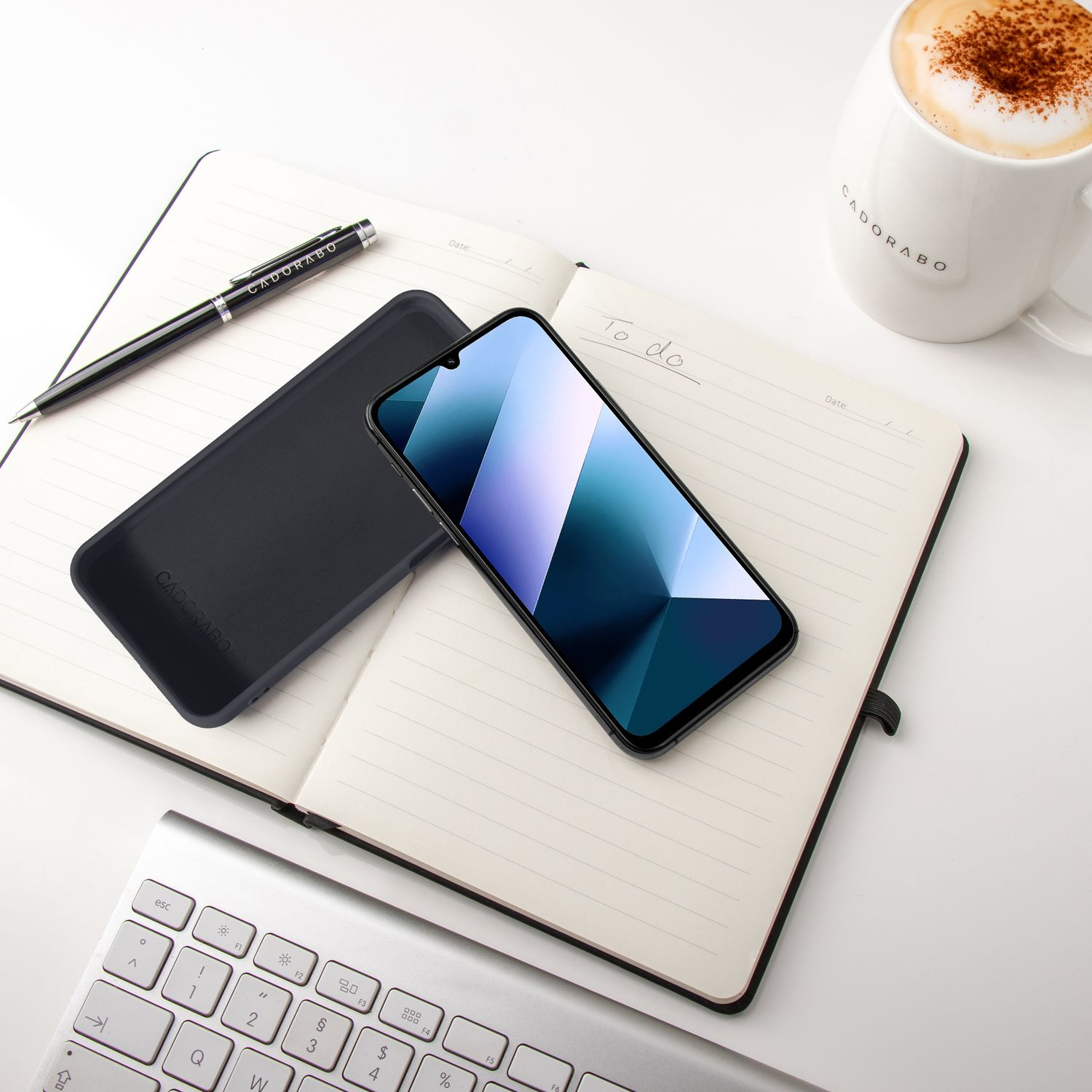 Notizbuch mit Handy, Stift, Tastatur und Kaffee auf weißem Tisch.