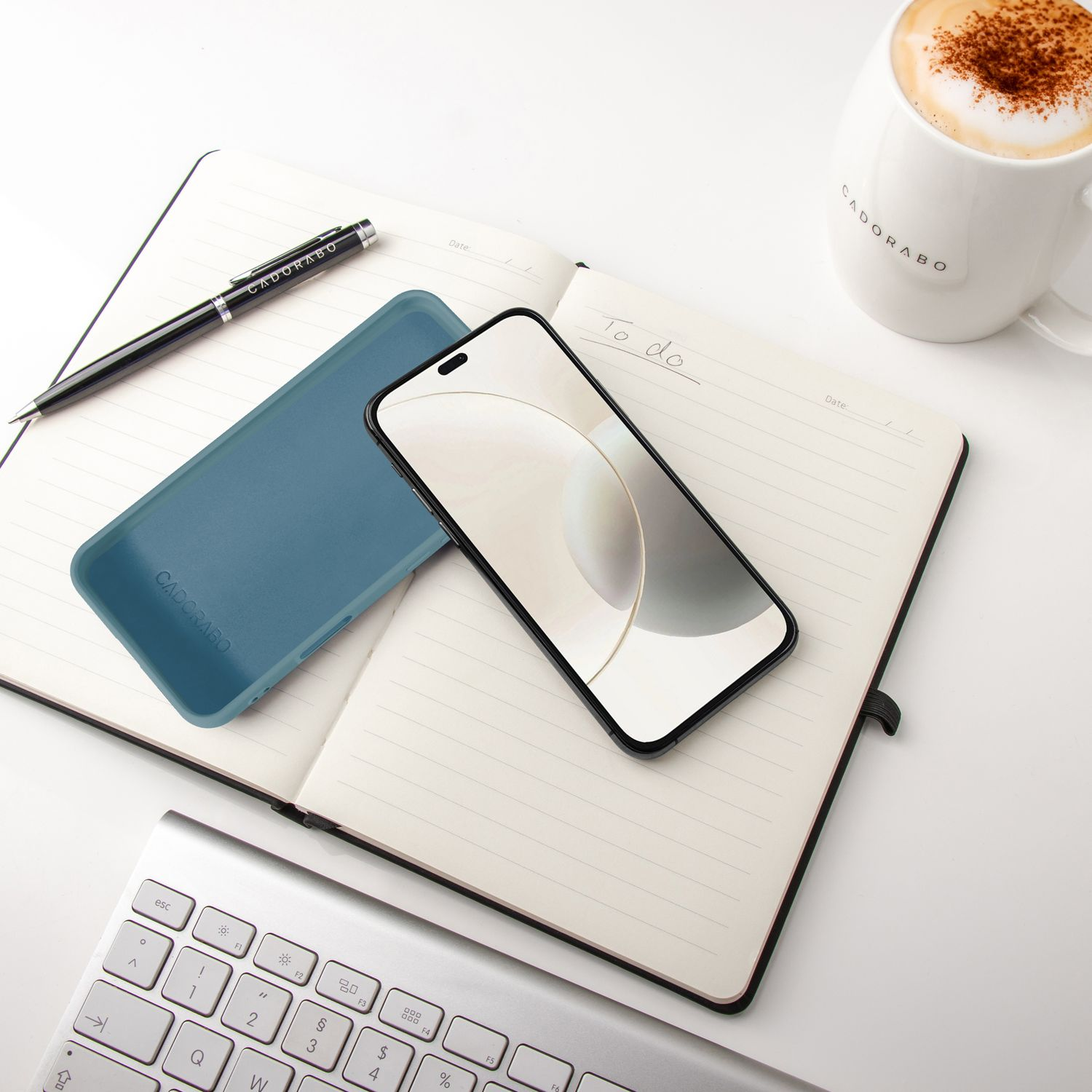 Ein Notizbuch mit Stift, Handy, Hülle und Kaffee auf einem weißen Tisch.