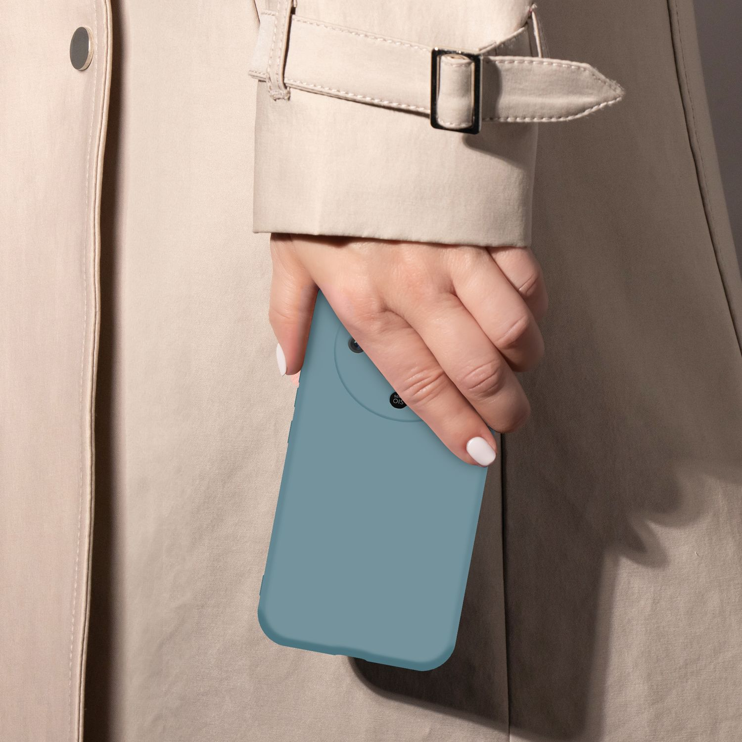 Eine Hand mit blauen Nägeln hält ein blaues Handy vor einem beigen Mantel.