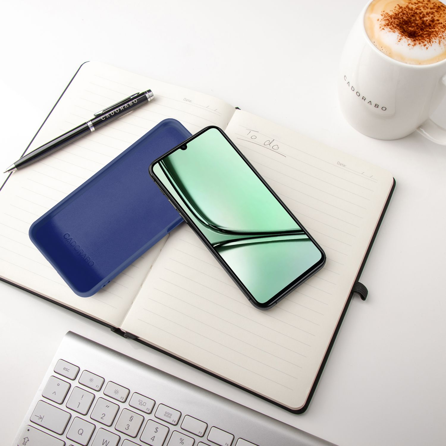 Ein Notizbuch mit Stift, Handy, Tastatur und Kaffee auf weißem Tisch.