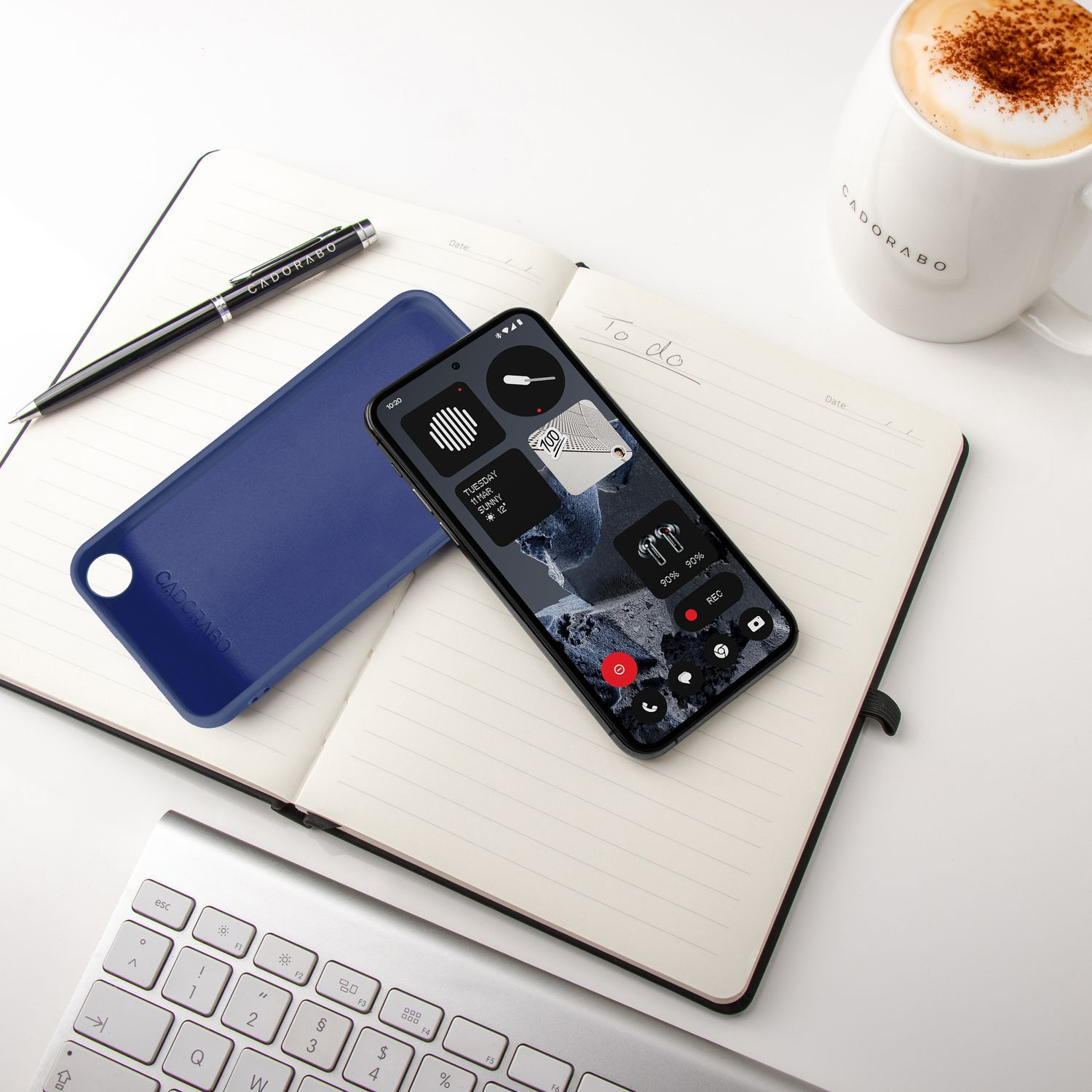Ein Smartphone liegt auf einem Notizbuch neben einer blauen Hülle und einer Tasse Kaffee.