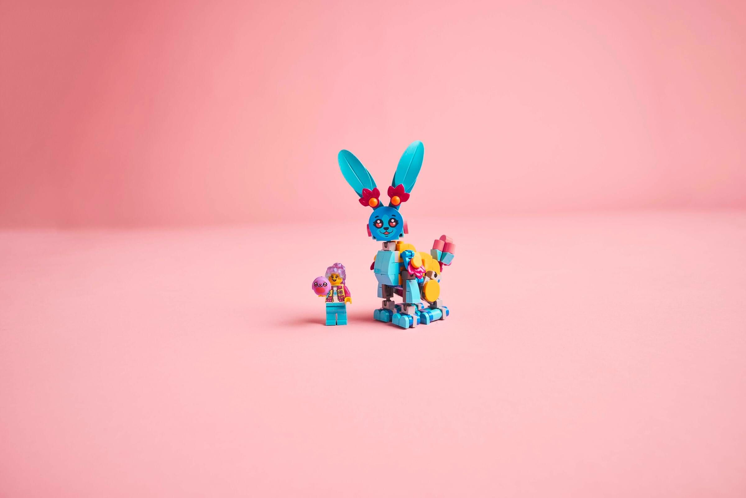 Eine Minifigur hält einen Ball neben einem blauen Lego-Hasen auf rosa Hintergrund.