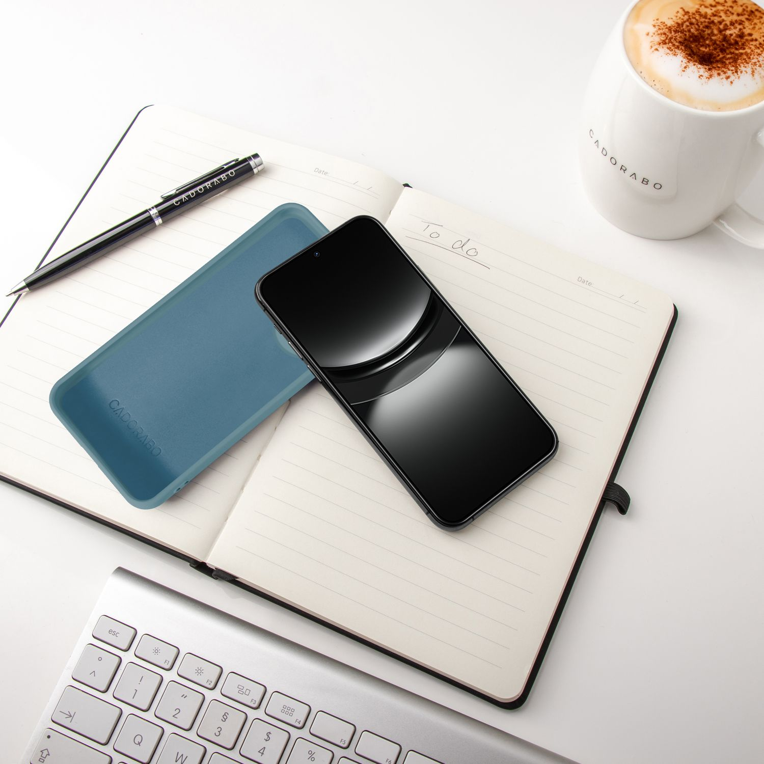 Handy, Notizbuch mit Stift, Hülle, Tastatur und Kaffee auf weißem Tisch.