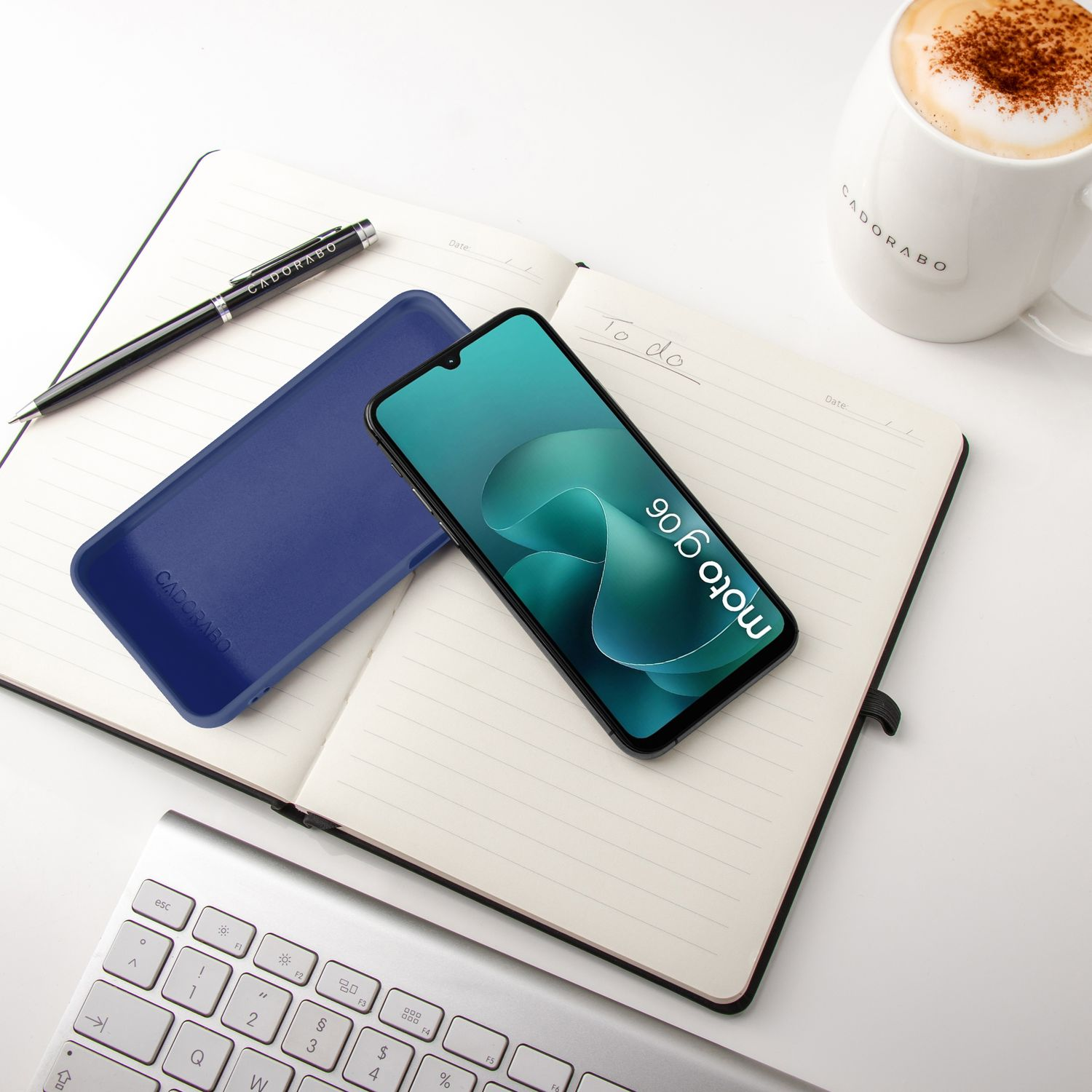 Ein Smartphone, eine Hülle, ein Notizbuch, eine Tasse Kaffee und eine Tastatur auf einem weißen Tisch.