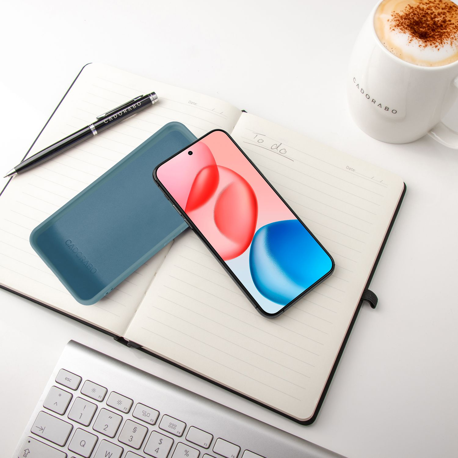 Ein Smartphone liegt auf einem Notizbuch neben einer Kaffeetasse und Tastatur.