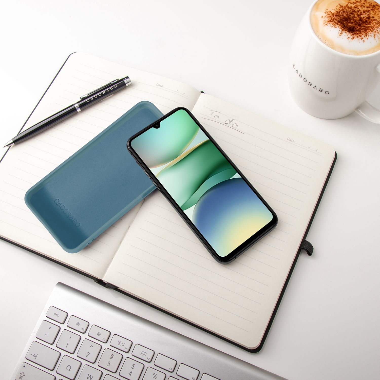 Ein Handy, Notizbuch, Stift, Tastatur und Kaffee auf einem weißen Schreibtisch.