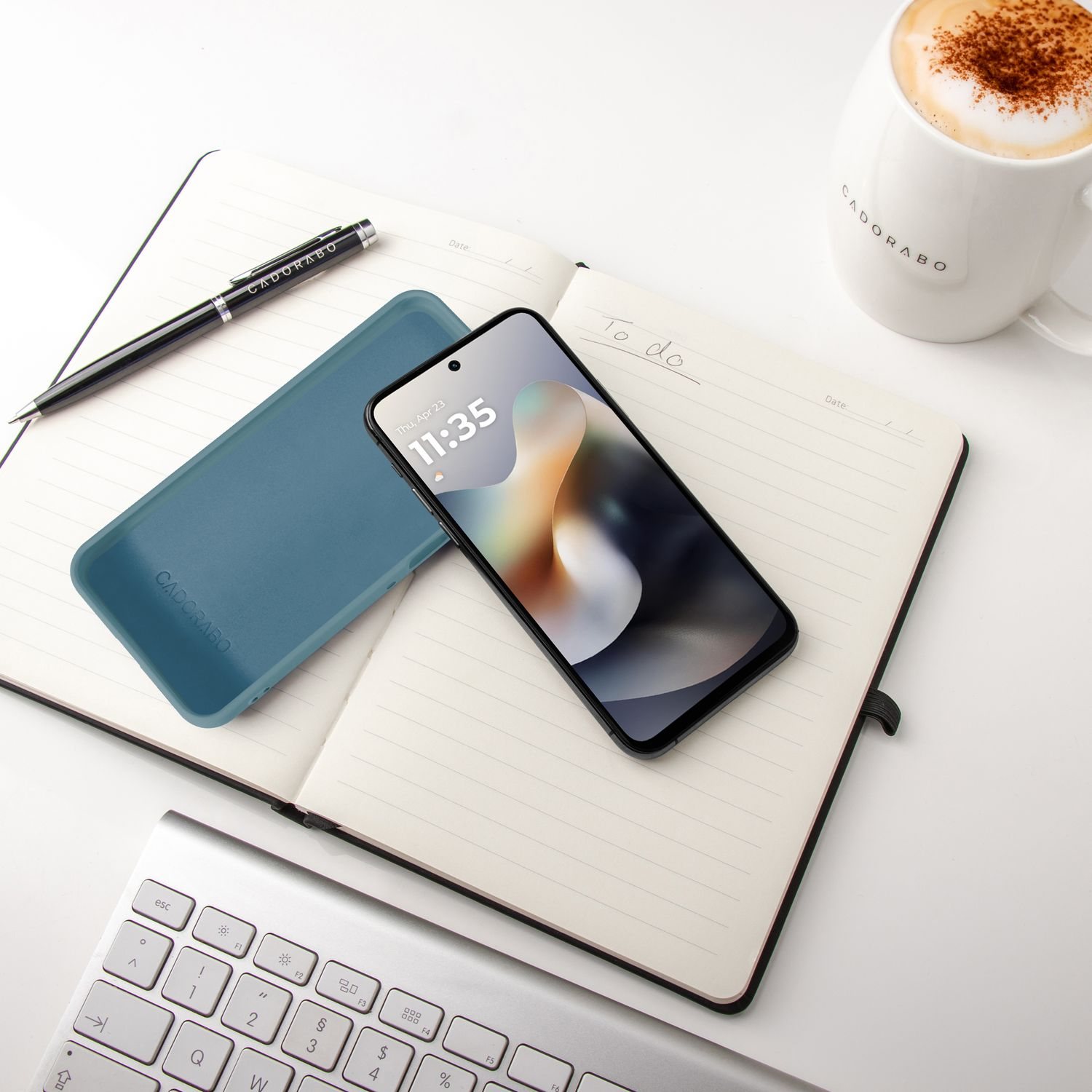 Ein Smartphone, Notizbuch, Stift, Tastatur und Kaffee auf weißem Tisch.