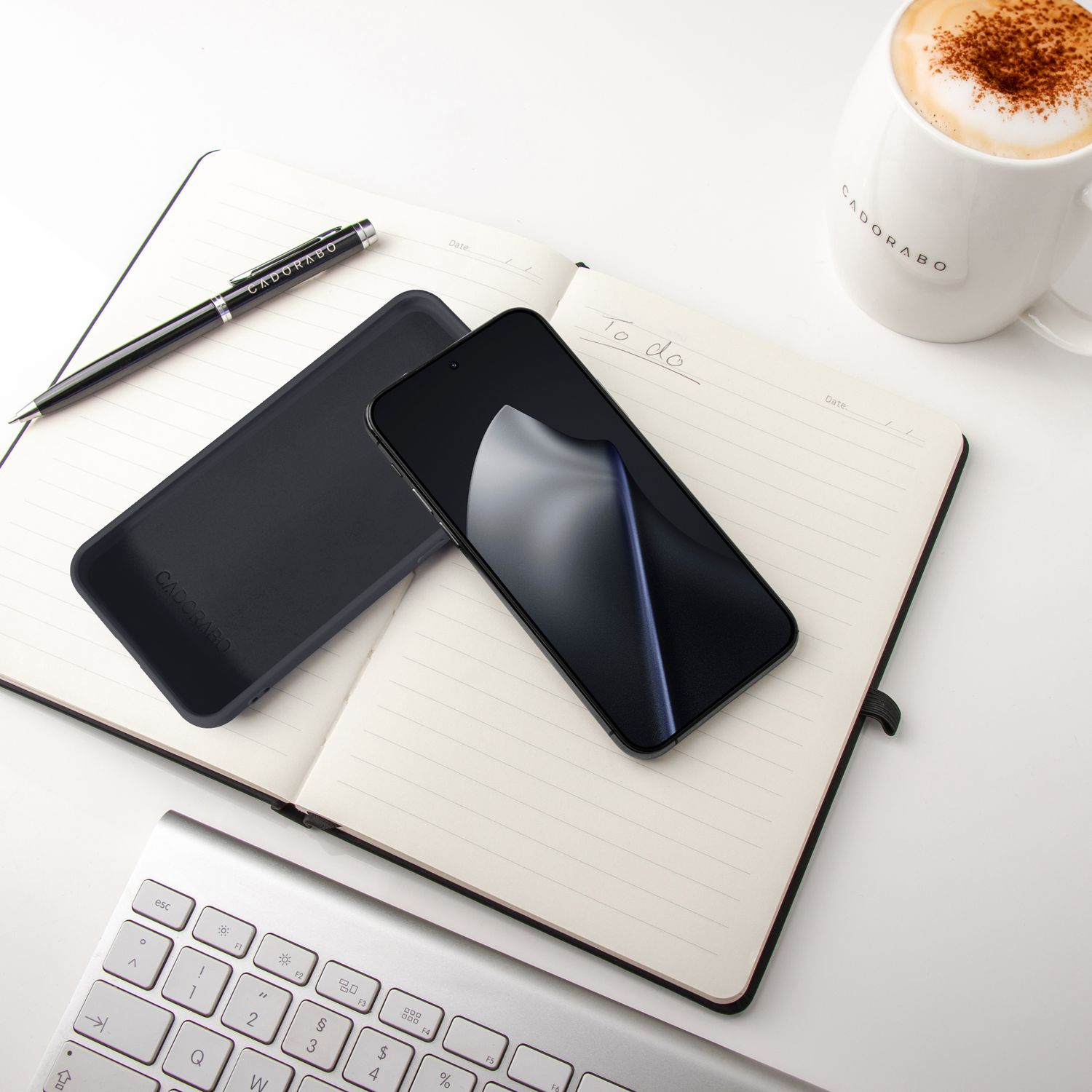 Ein schwarzes Smartphone, Hülle, Notizbuch, Stift und eine Kaffeetasse liegen auf einem Schreibtisch.