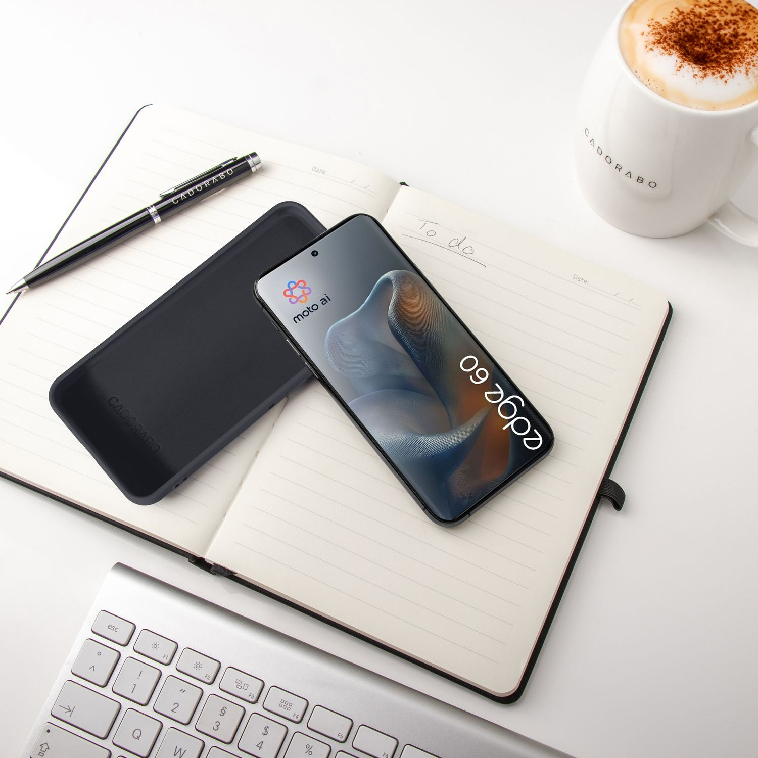 Ein schwarzes Handy, Hülle, Stift, Notizbuch, Tastatur und Kaffeetasse auf weißer Fläche.