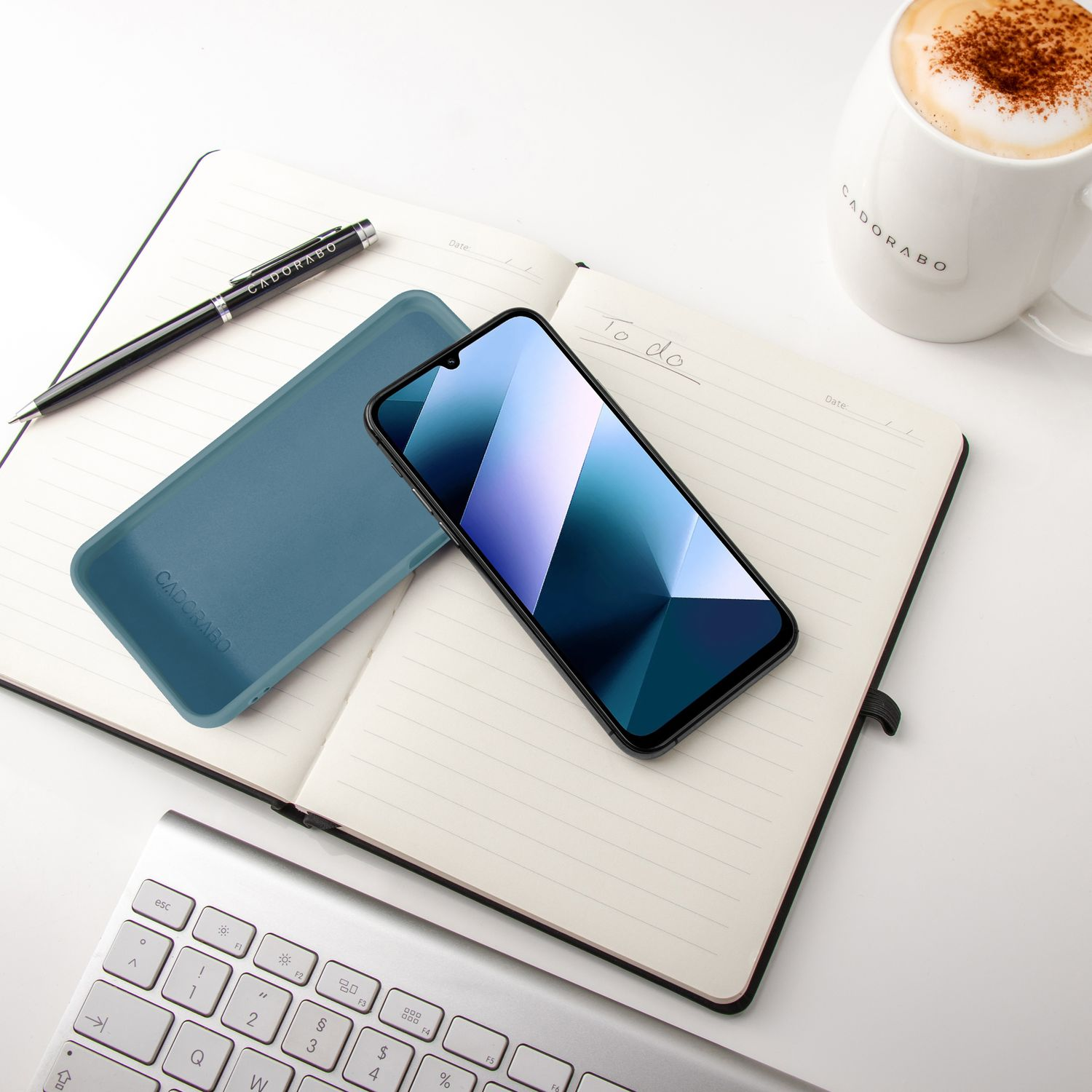 Ein Notizbuch mit Stift, Handy, Tastatur und Kaffee auf weißem Tisch.