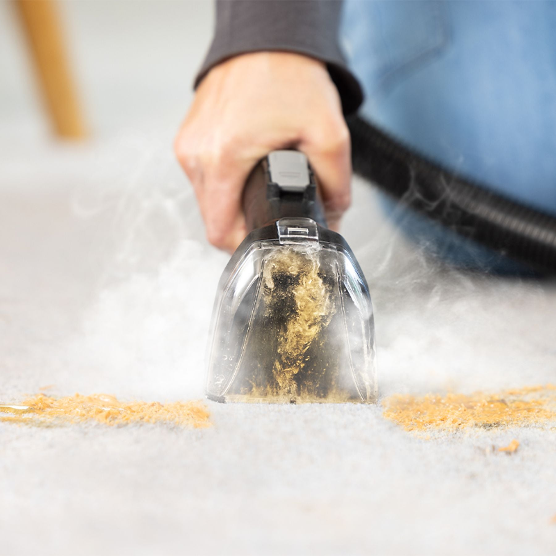 A hand uses a wet vacuum to clean dirt from a carpet. Steam is visible.