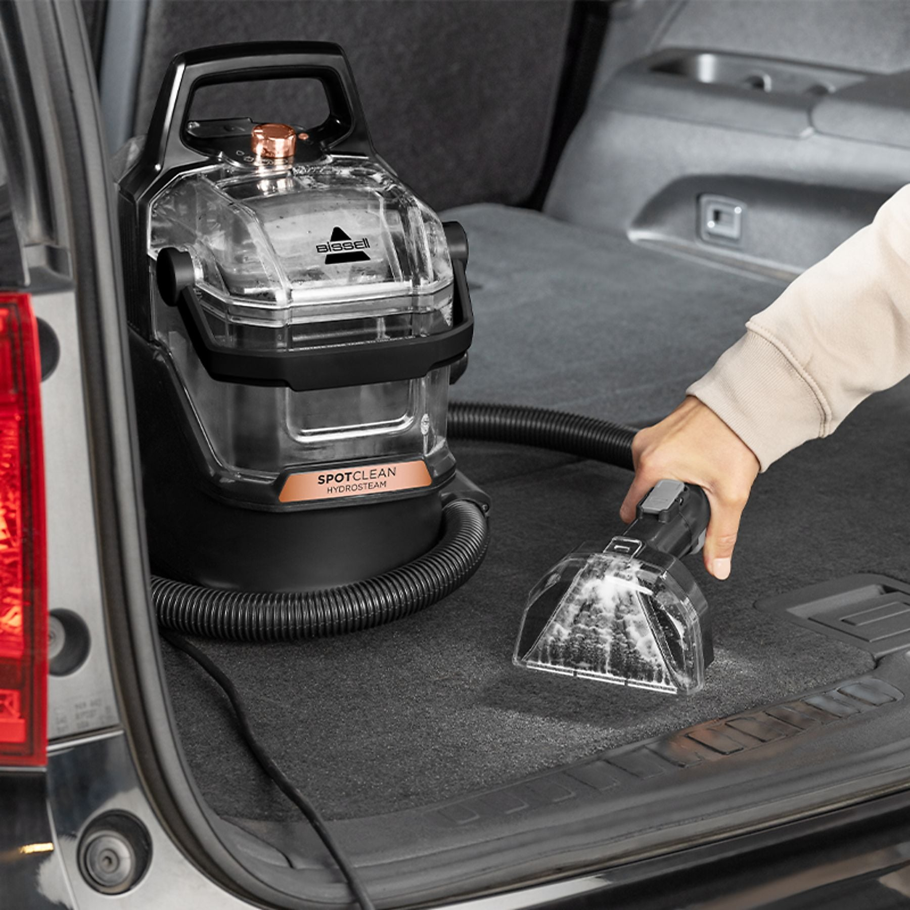 A handheld carpet cleaner is used on the floor of a car's trunk.