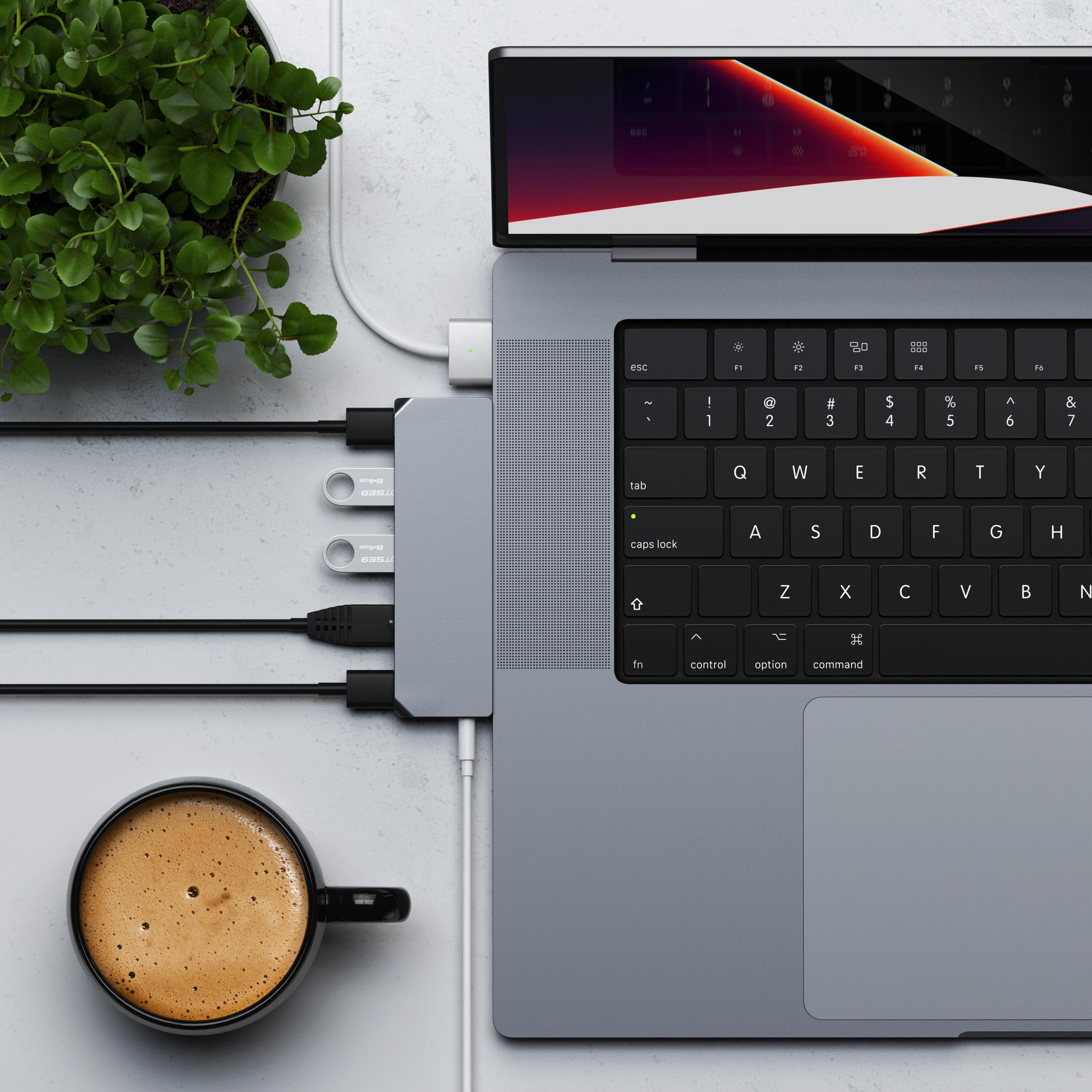 Un laptop con hub, due chiavette USB, una tazza di caffè e una pianta.