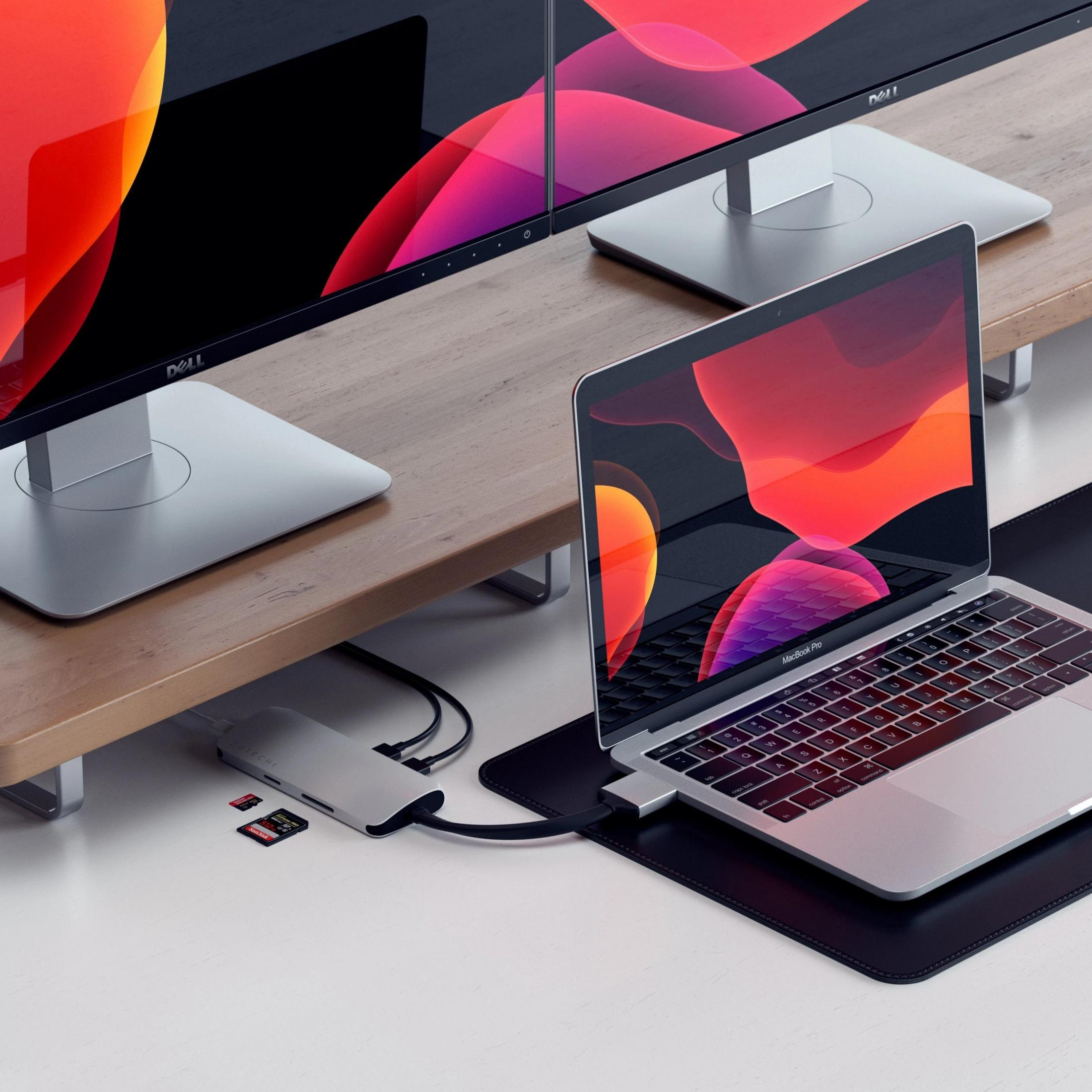 Un MacBook Pro e due monitor Dell su un supporto in legno con hub e schede SD.