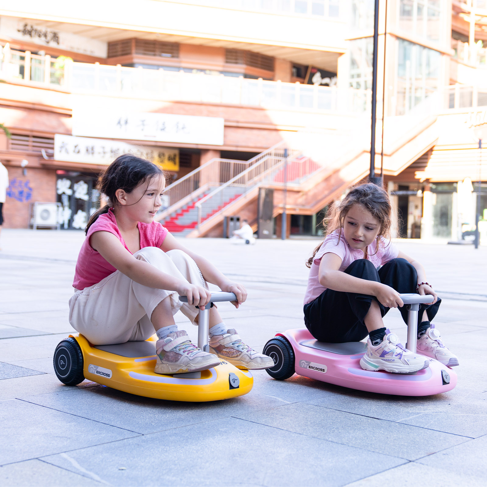 Zwei Mädchen fahren Elektroroller. Einer gelb, einer pink, auf einem gepflasterten Platz.
