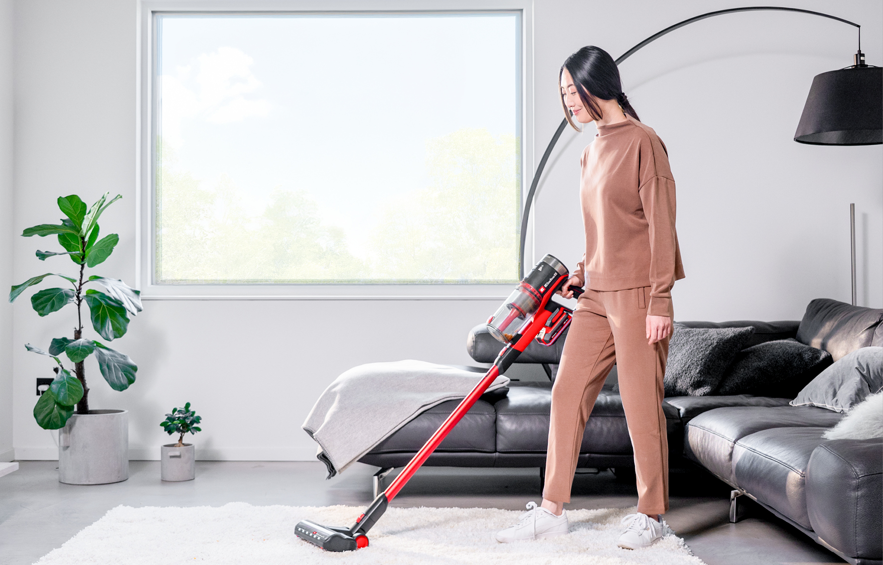 Frau saugt Teppich mit rotem Akku-Staubsauger. Wohnzimmer mit Sofa und Pflanzen.