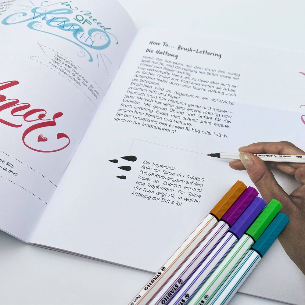 Una mano tiene una penna su un libro di brush lettering, con pennarelli colorati accanto.