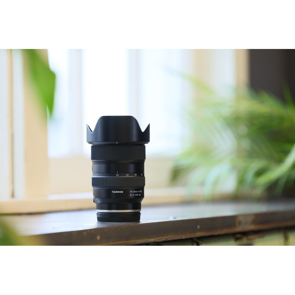 Tamron lens met lenskap op een plank, planten op achtergrond.