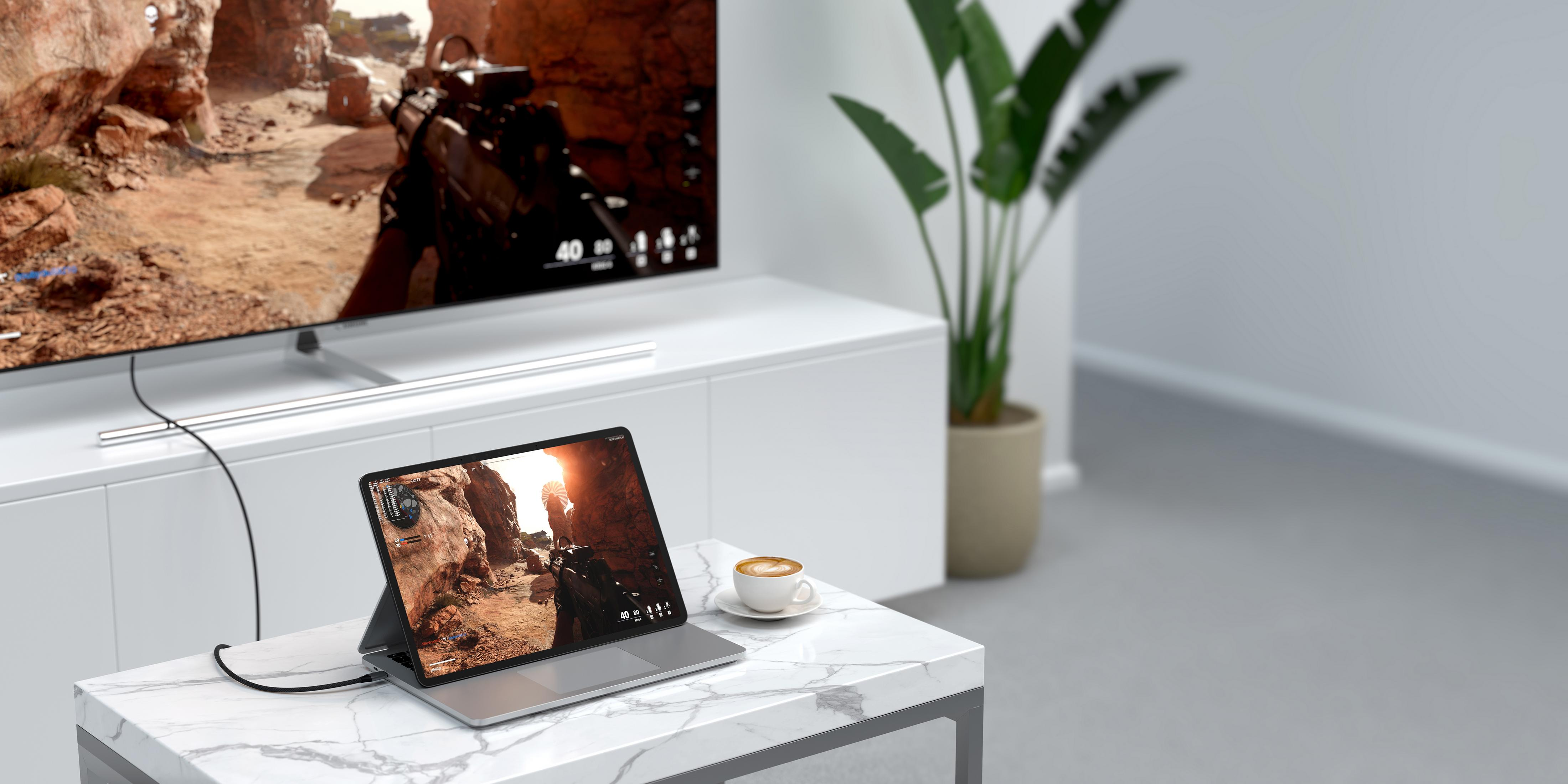 Ein Laptop mit einem Videospiel, daneben eine Kaffeetasse, im Hintergrund ein Fernseher.