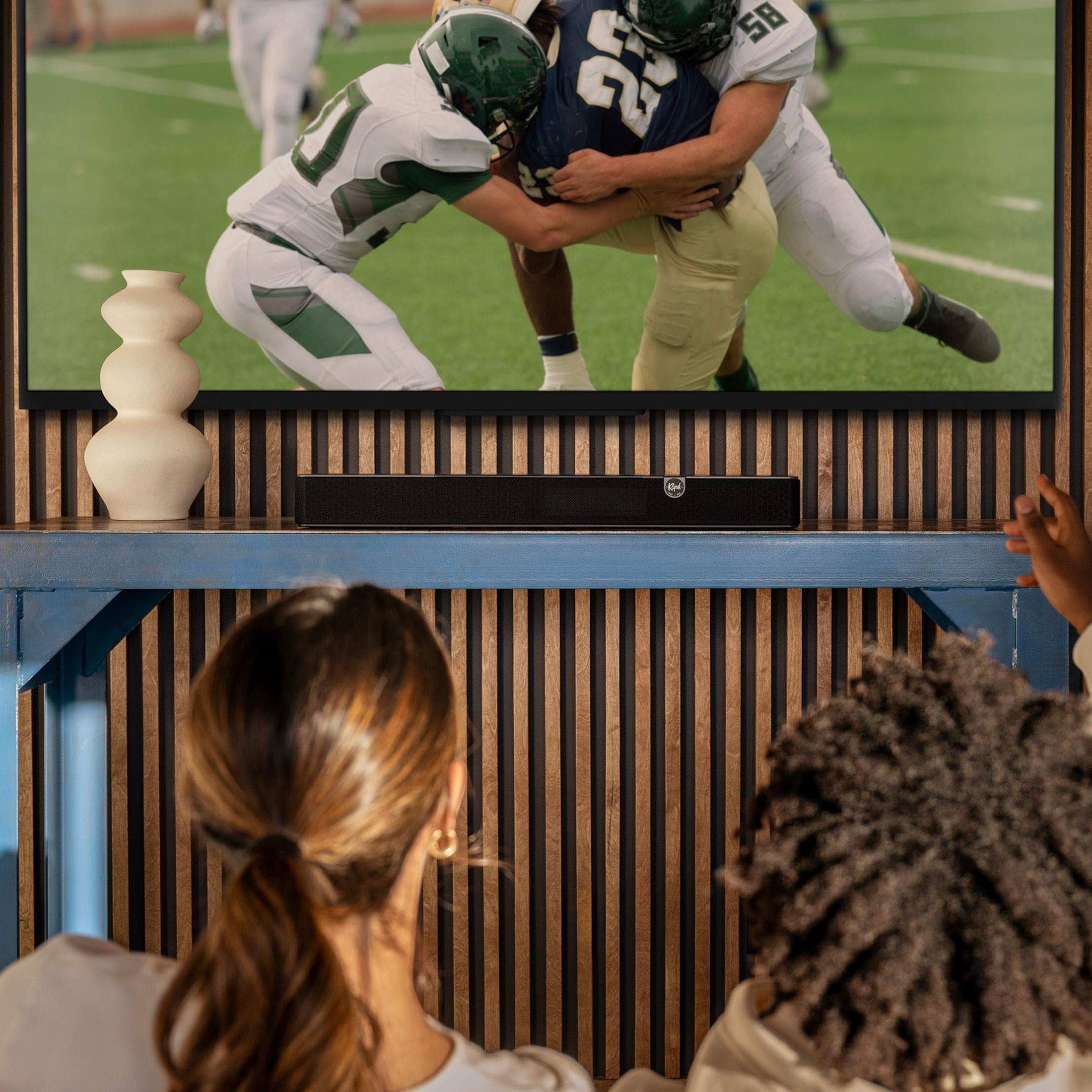 Dwie osoby oglądają futbol w telewizji. Poniżej soundbar.