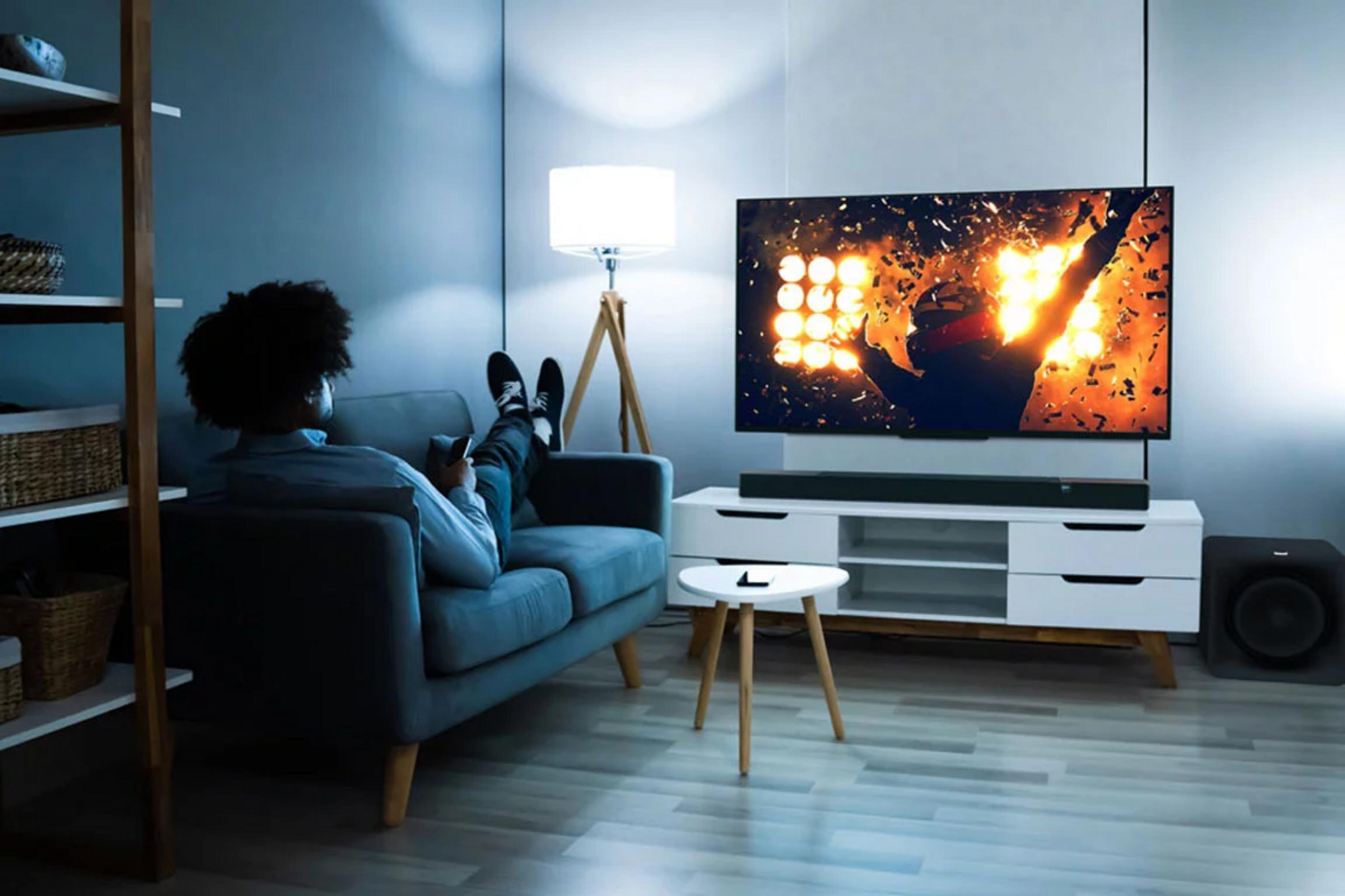 Mężczyzna na kanapie ogląda TV z fajerwerkami. Lampa i półka widoczne.