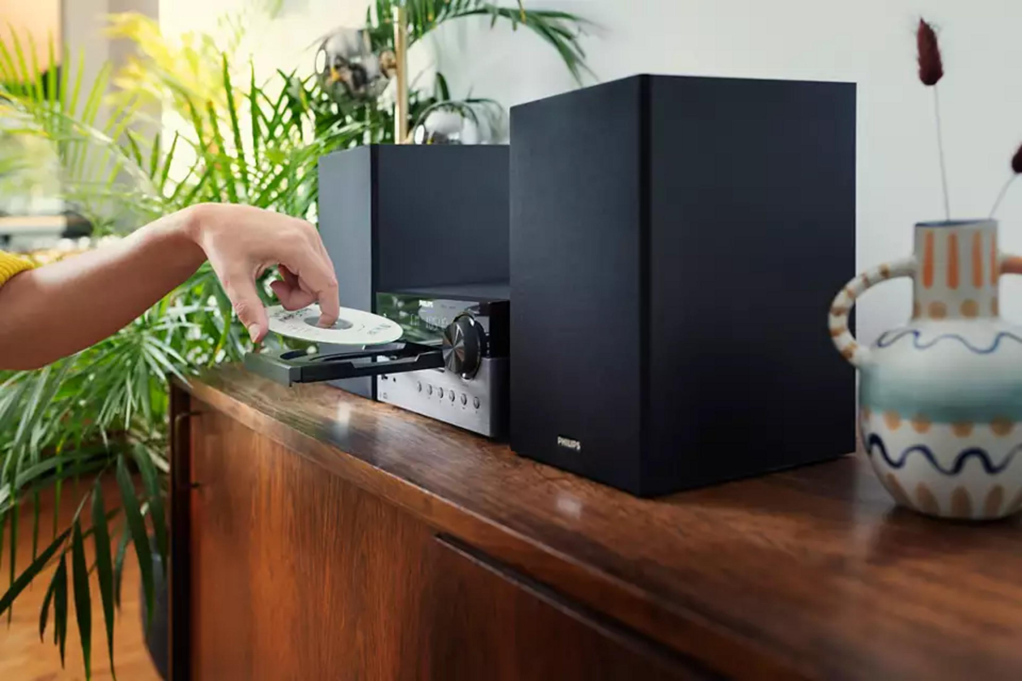 Ręka wkłada płytę CD do wieży stereo. Po lewej rośliny, po prawej wazon.