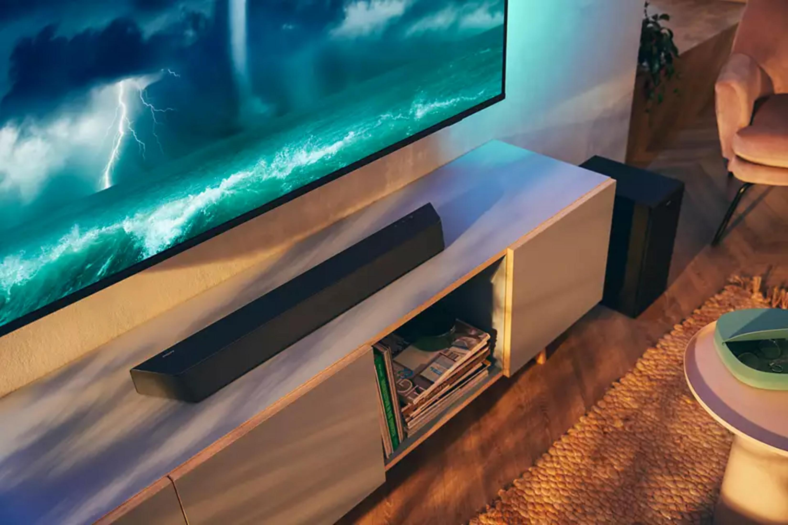 Czarny soundbar na szarej szafce RTV przed dużym ekranem telewizora.