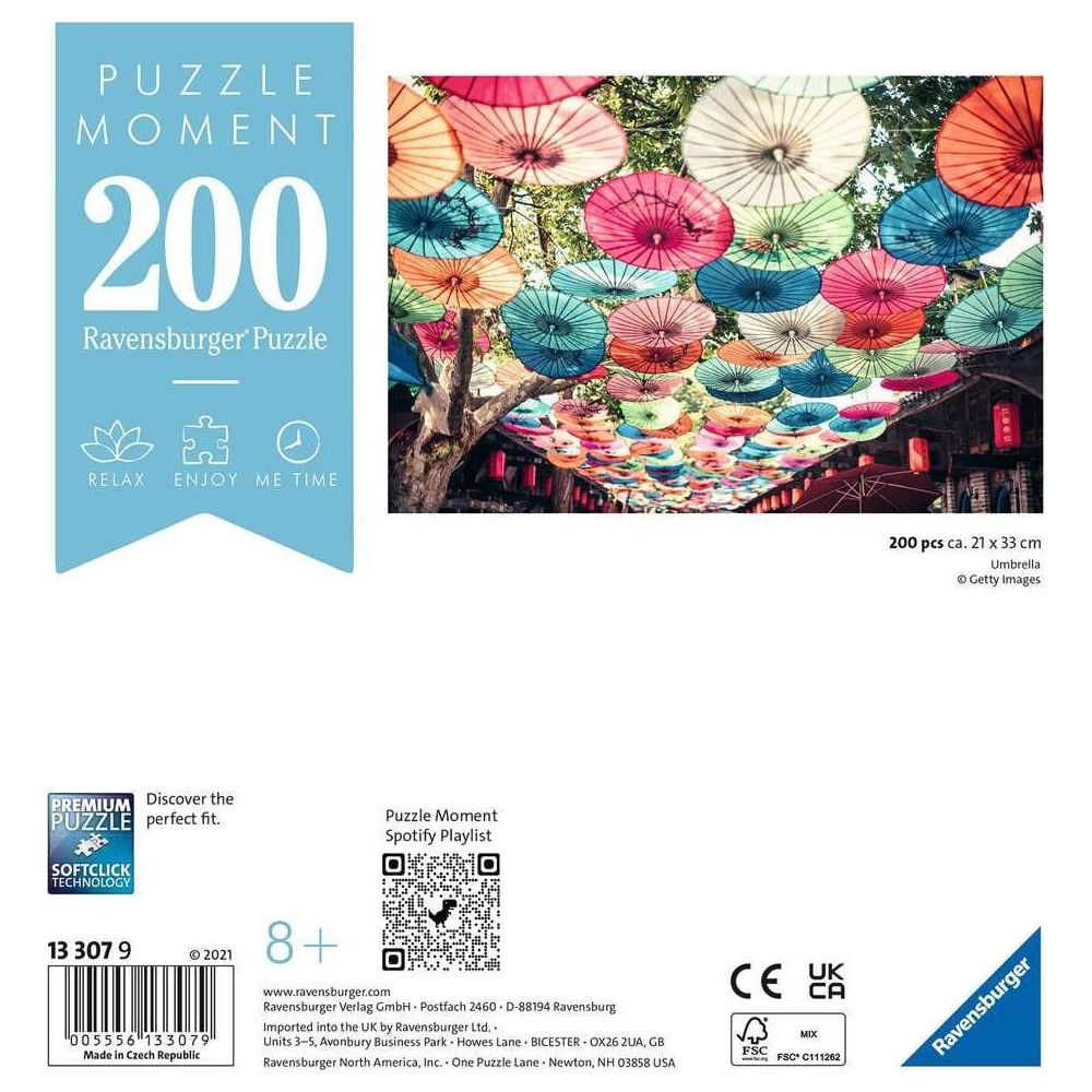 Sfondo: Molti ombrelli colorati pendono su una strada, primo piano confezione puzzle Ravensburger.